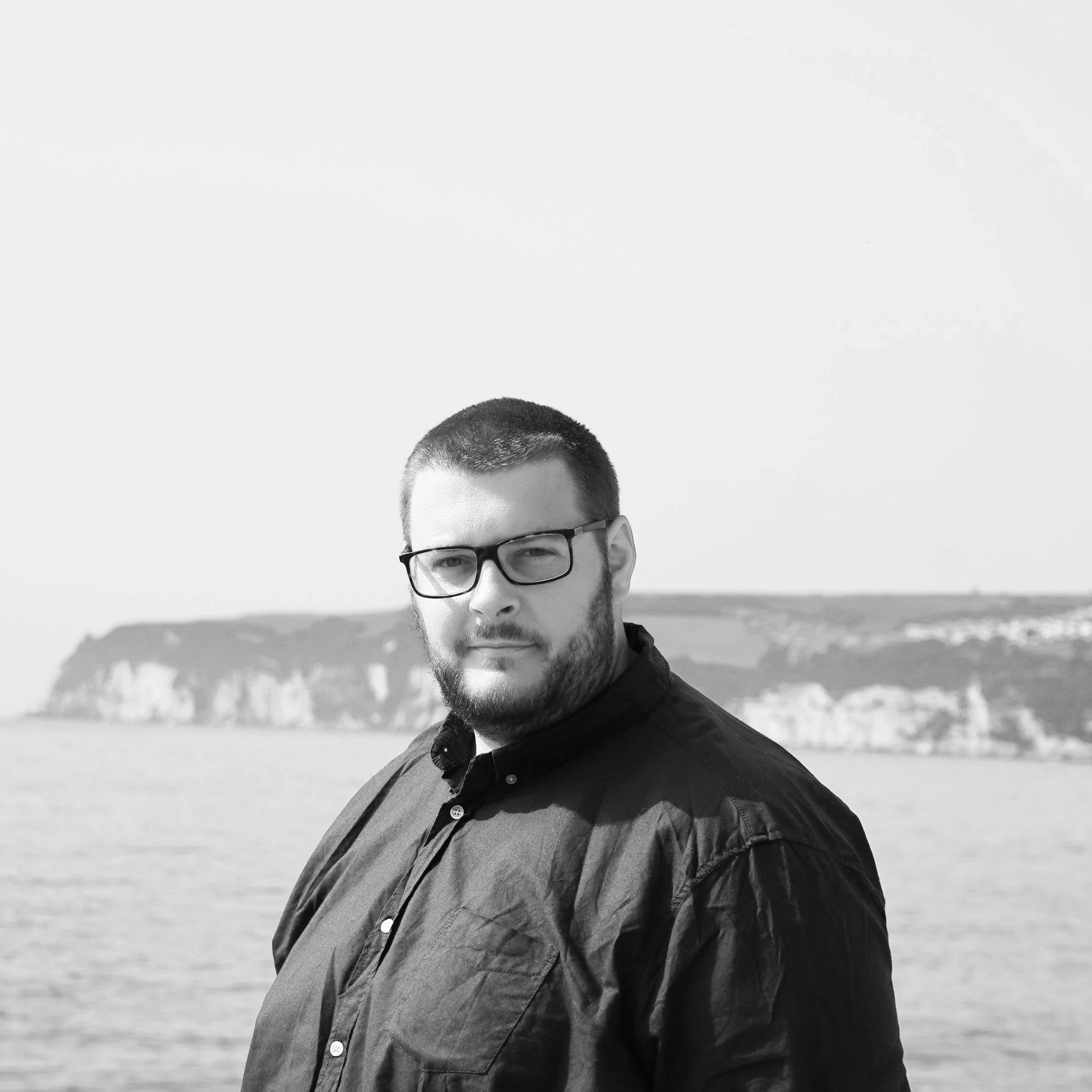 Black and white photograph of a man with glasses and a beard, standing outdoors near water with cliffs in the distance.