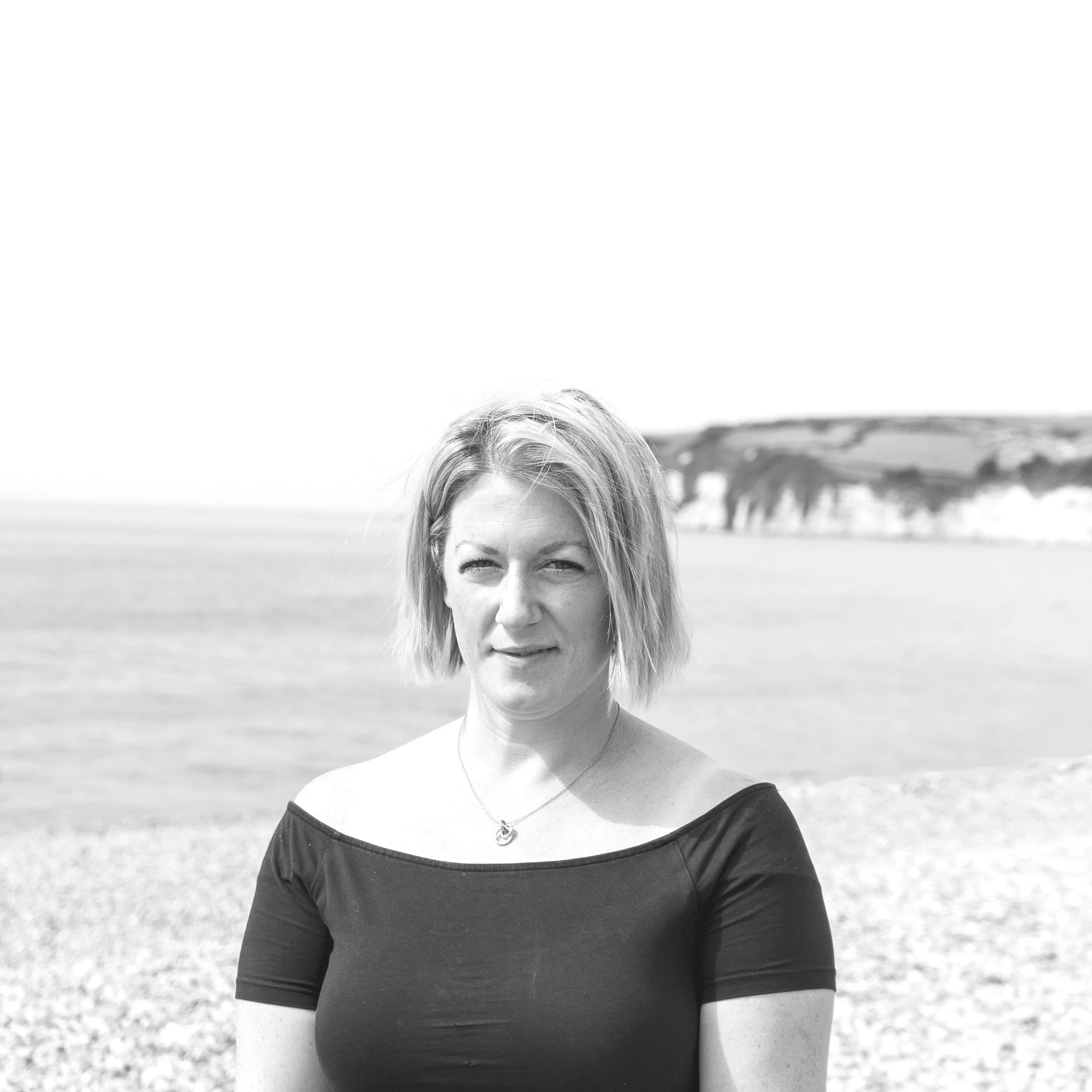 Black and white photo of a woman with short blonde hair standing on a beach with water and cliffs in the background.