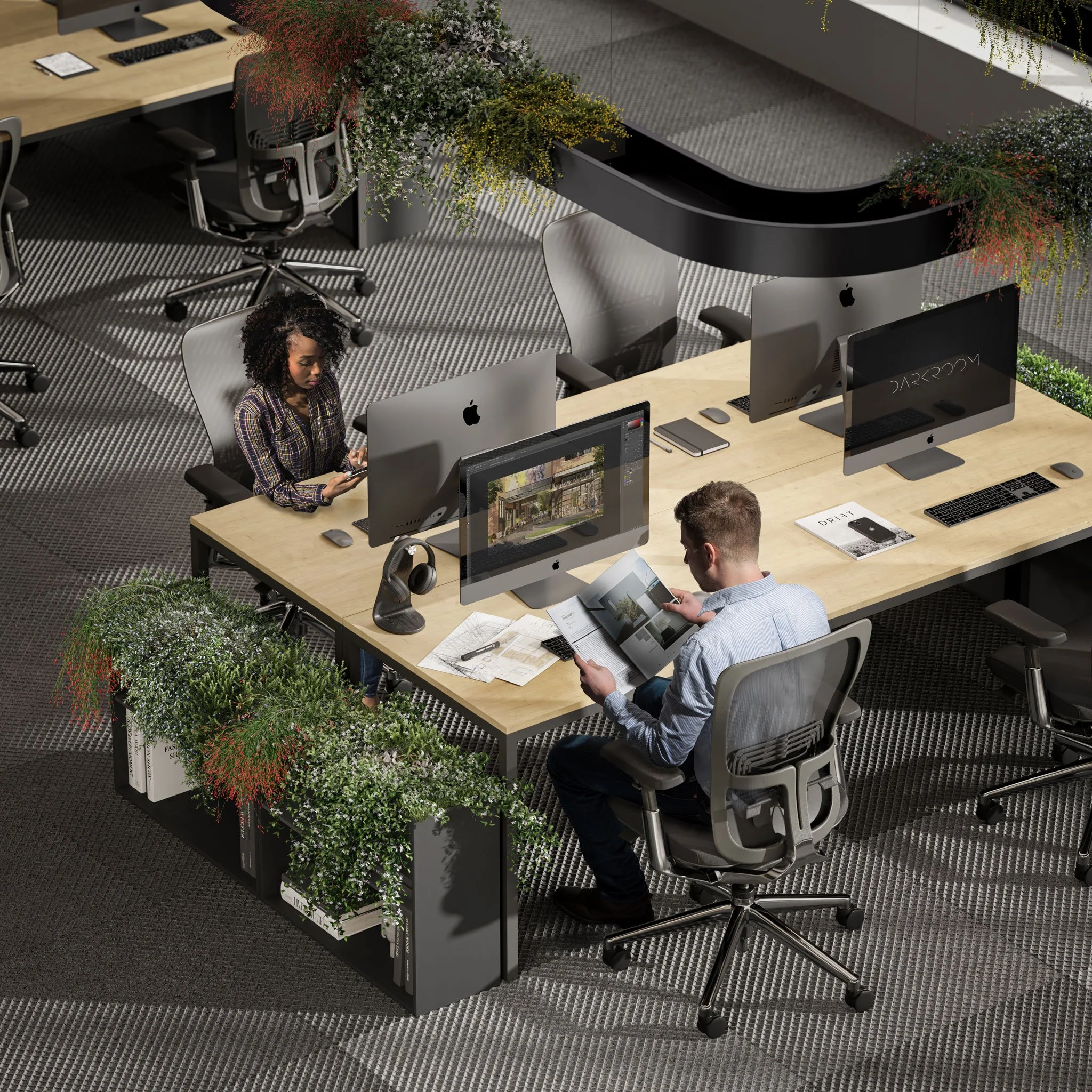 Office workspace with two people working at a shared desk, surrounded by potted plants, with multiple Apple computers and office supplies.