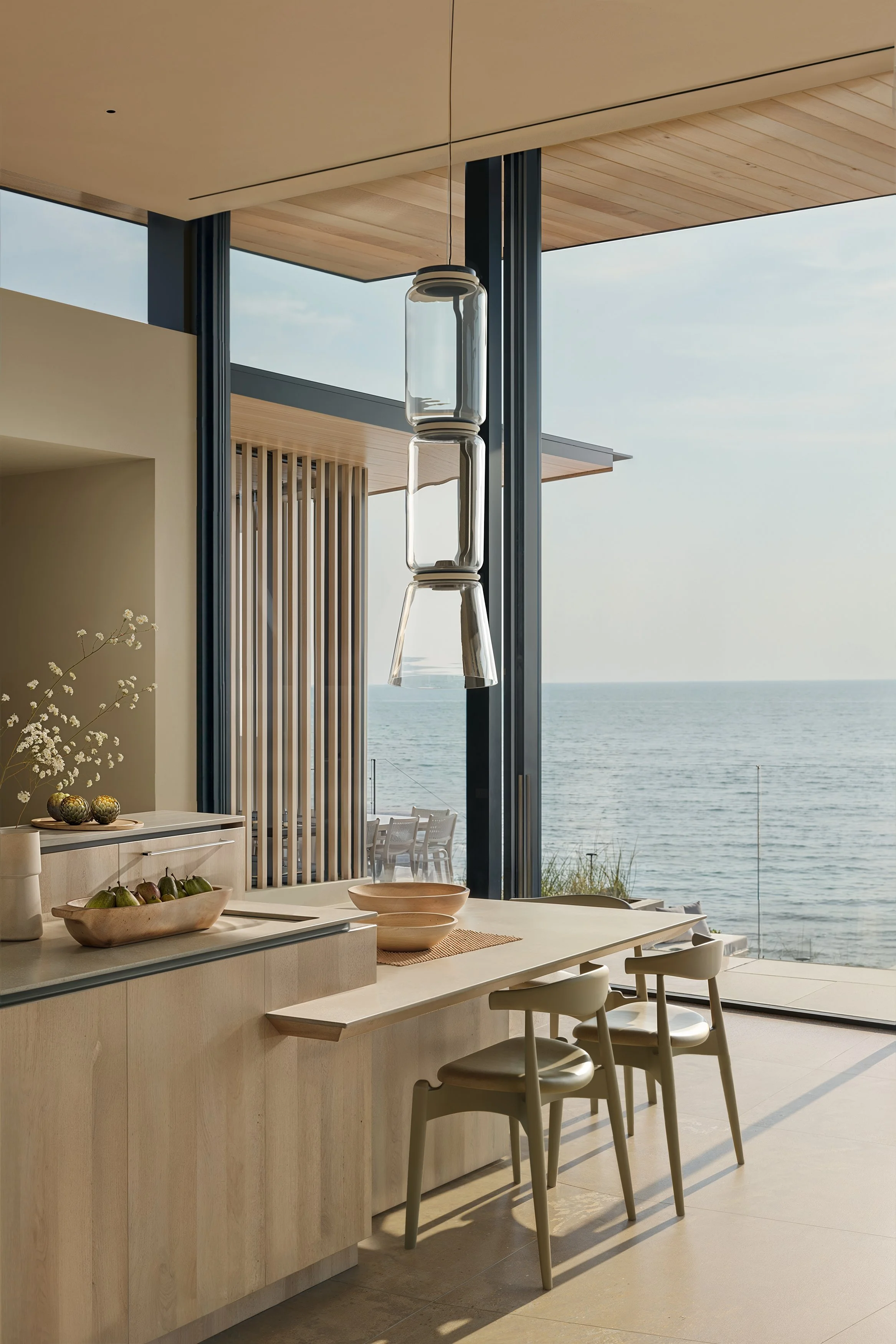 Modern kitchen with a view of the ocean, featuring wooden cabinets, a hanging glass pendant light, and beige chairs near a counter.