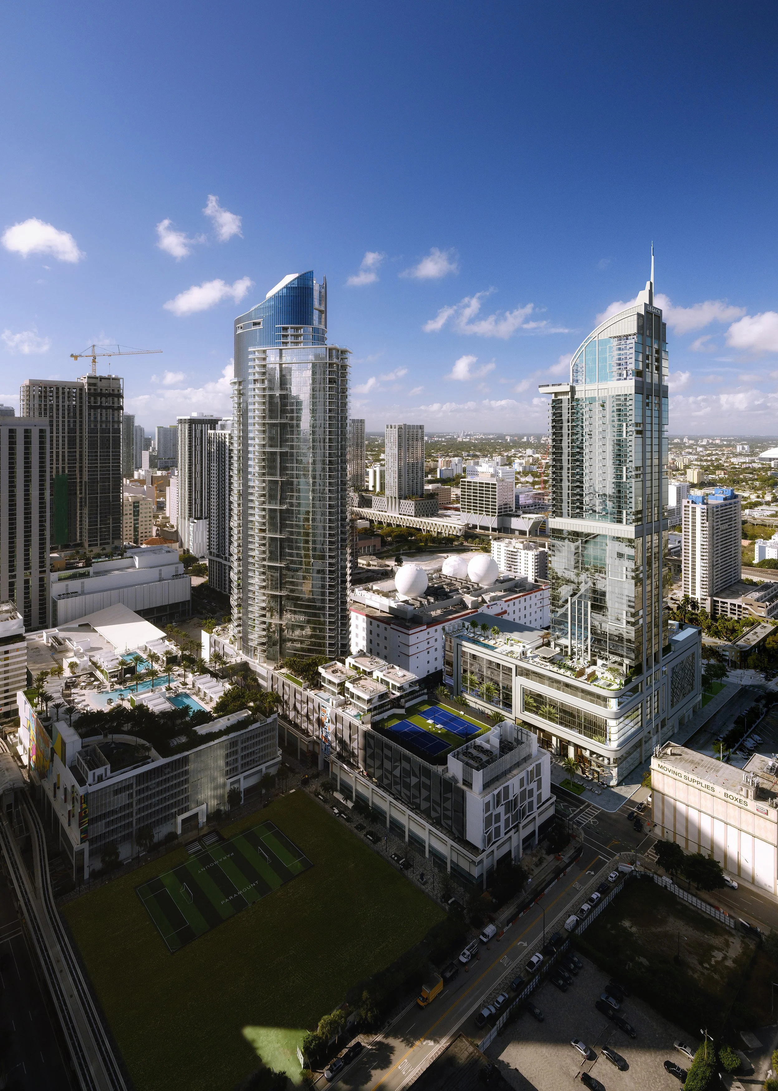 Aerial view of a city skyline featuring tall modern glass skyscrapers, a swimming pool, tennis court, and a football field, with a clear blue sky overhead.