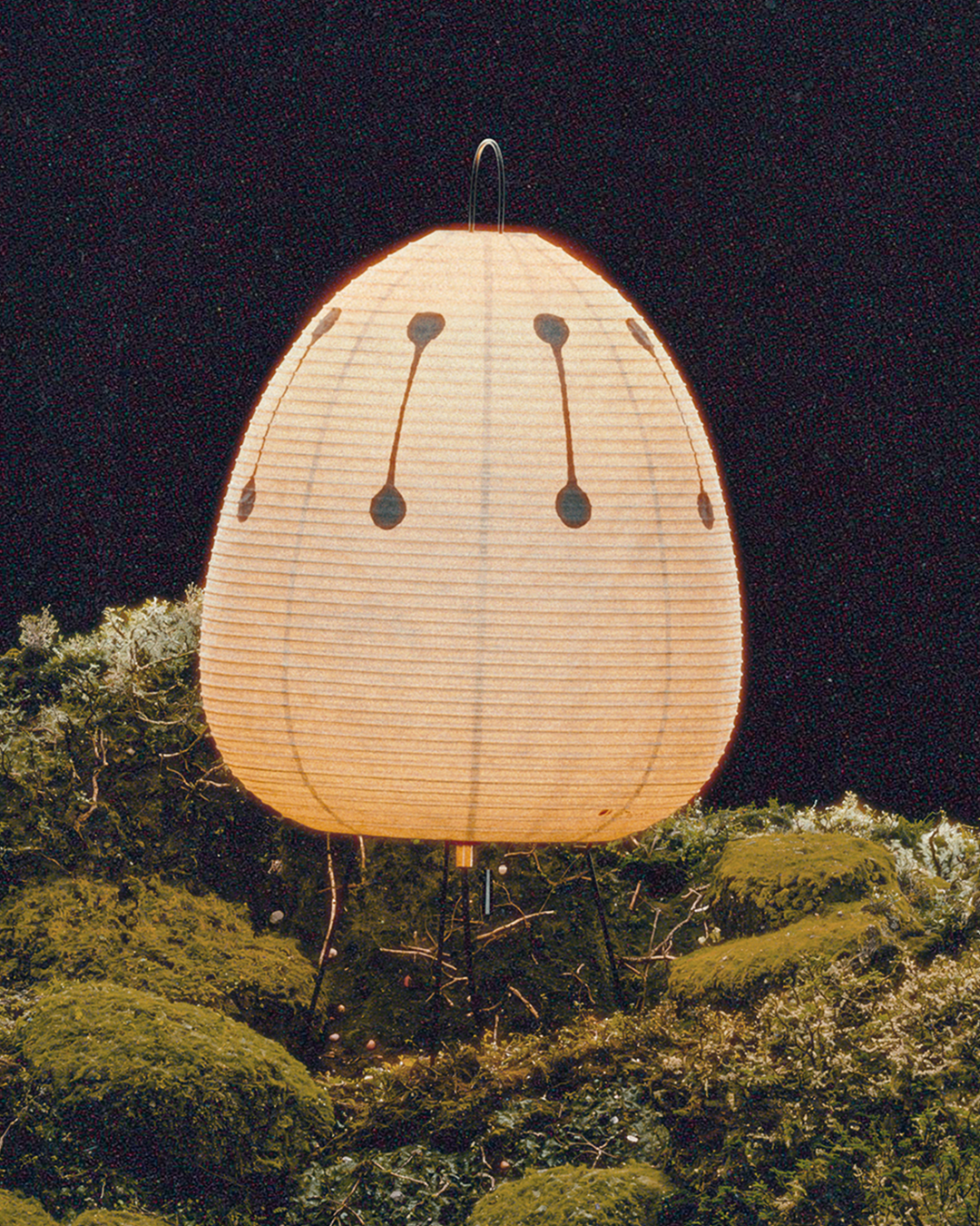 A large, egg-shaped paper lantern with painted face features, placed outdoors at night over a bed of moss and foliage. ISAMU NOGUCHI AKARI