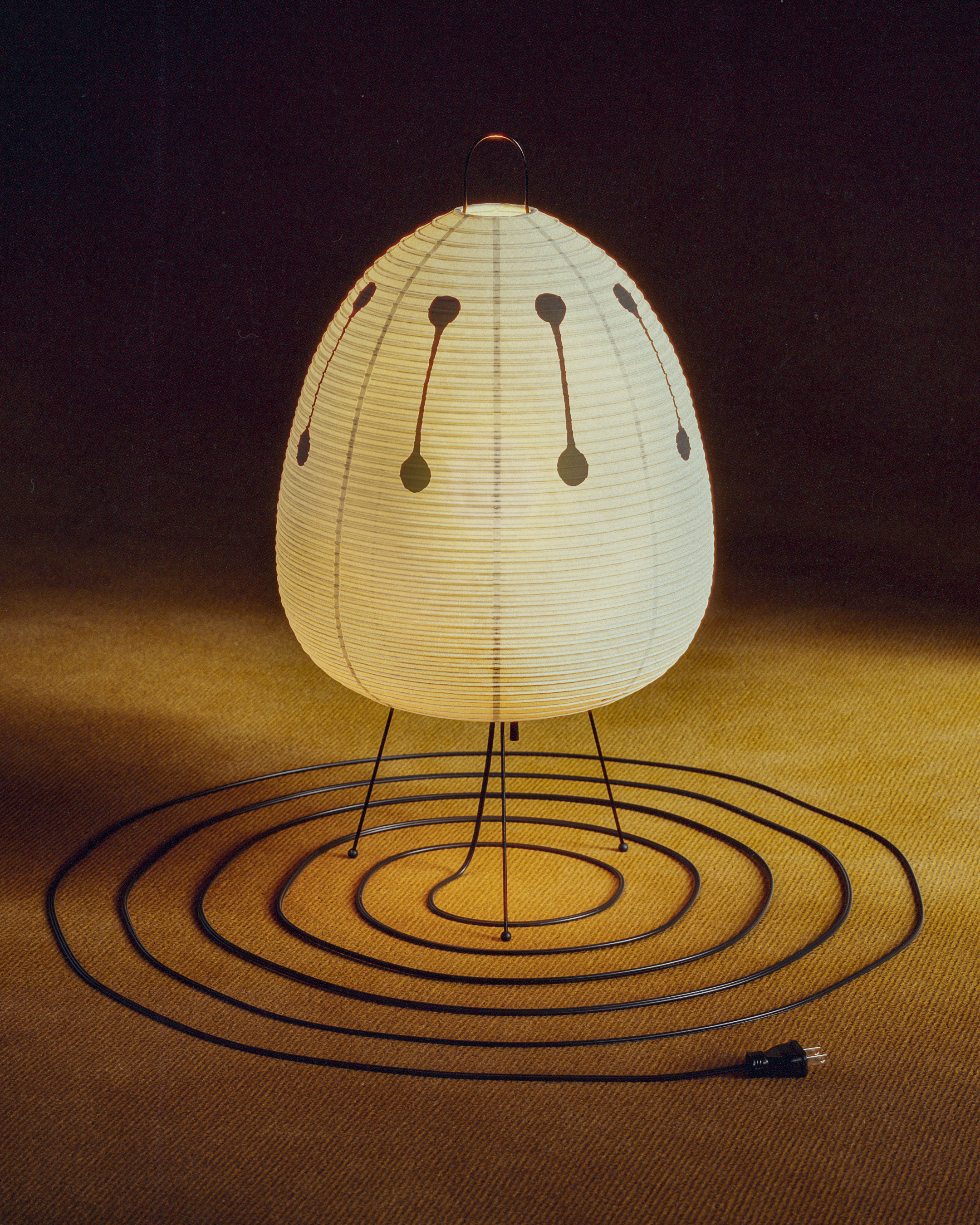 An Akari light sculpture by Isamu Noguchi softly glows as a paper lantern on slender metal legs, casting warm light onto a textured floor with a coiled power cable.