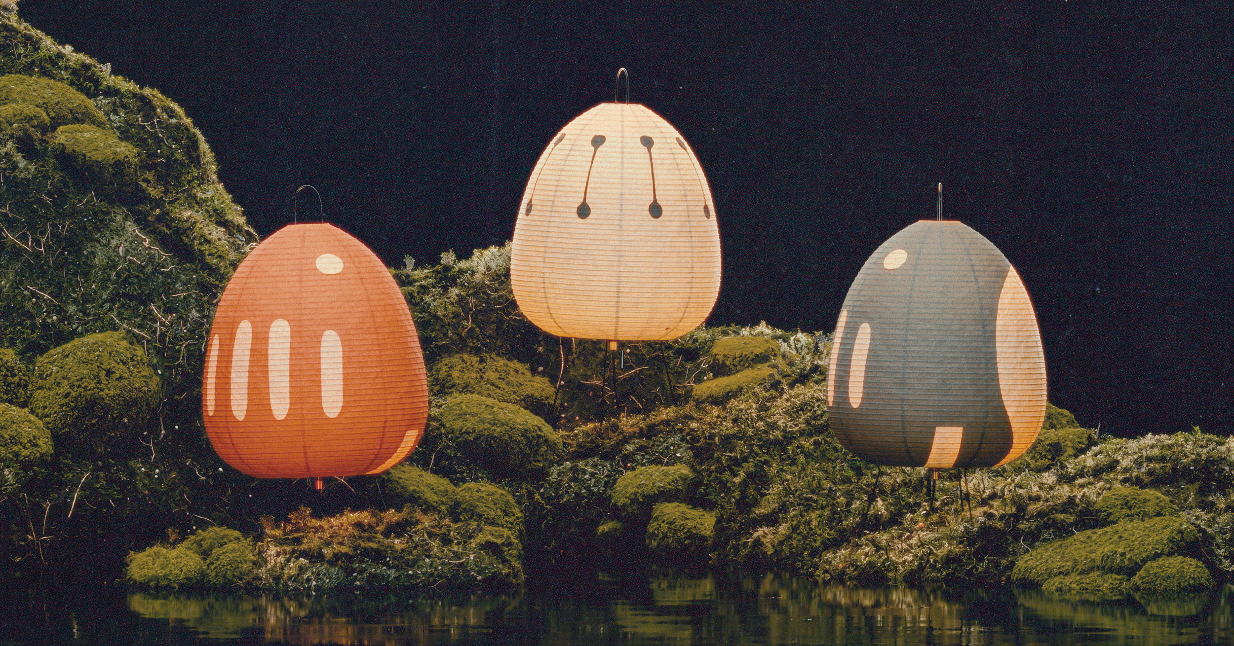 Three paper lanterns hanging above moss-covered rocks and water at night. 	
ISAMU NOGUCHI AKARI 
