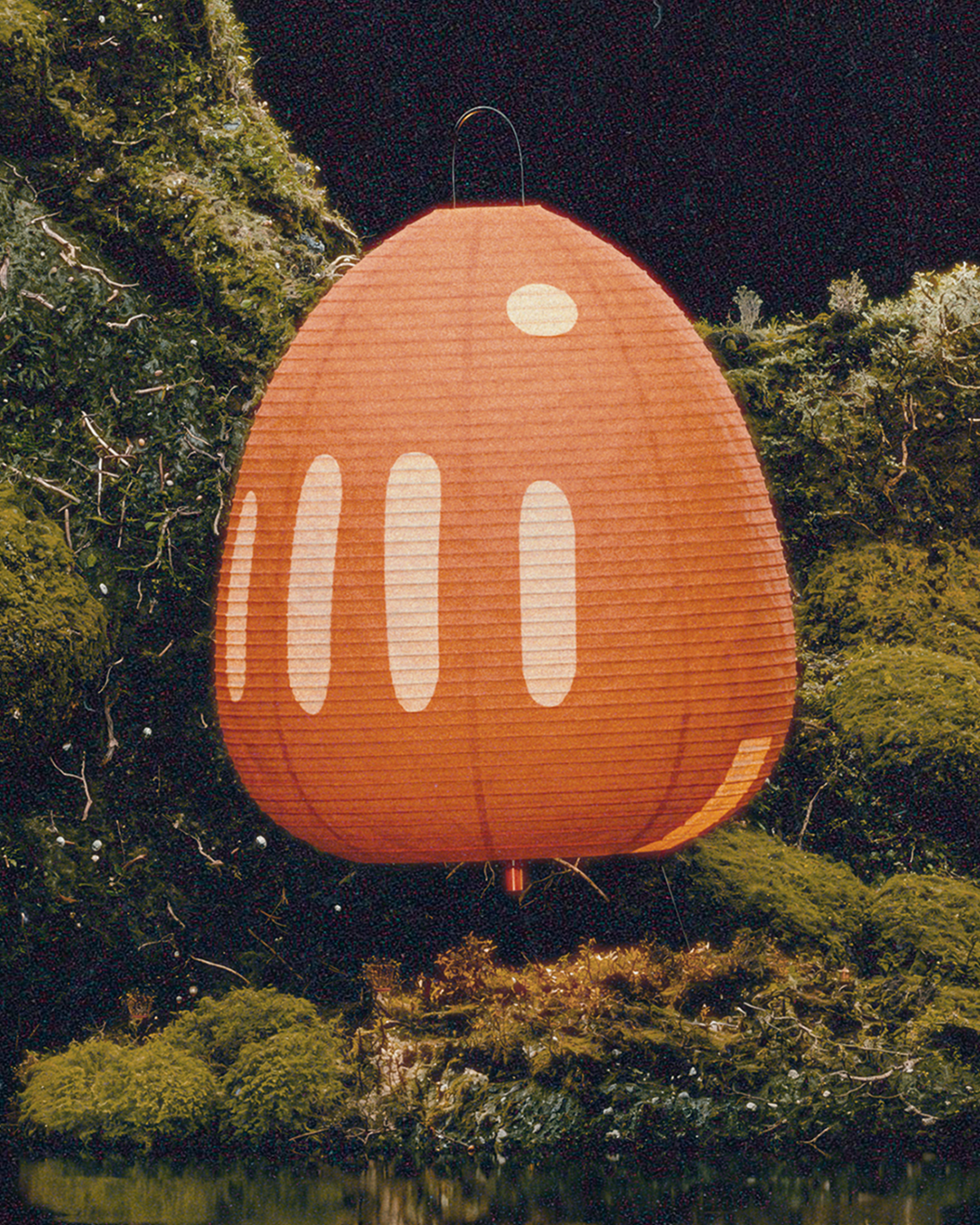 A large red paper lantern with white details, hanging among green foliage at night. ISAMU NOGUCHI AKARI