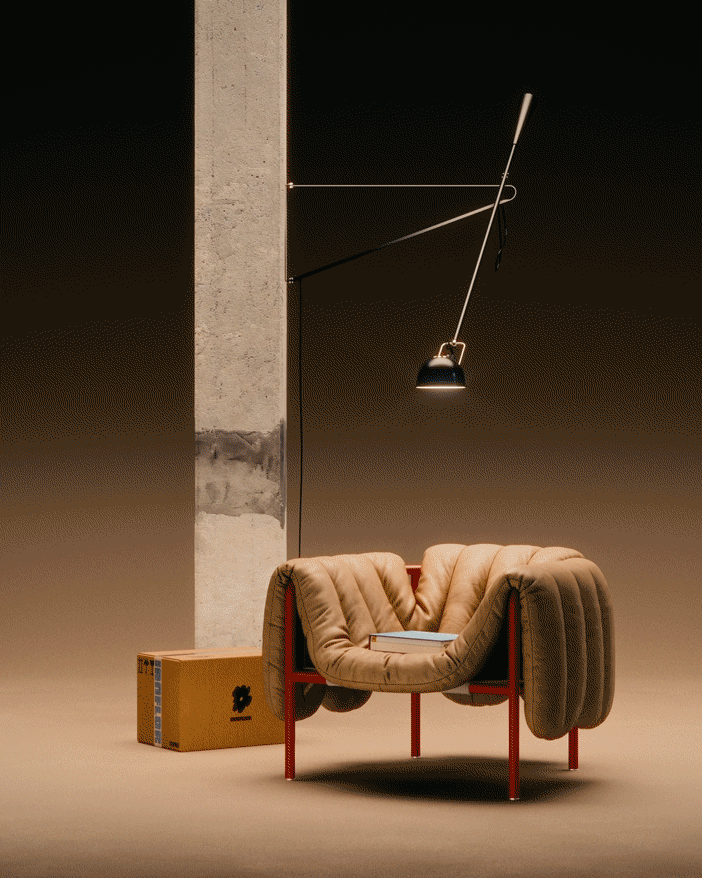 A modern beige armchair with red metal legs placed in a minimalist space, with a book resting on it, next to a wooden box and a tall concrete pillar. PUFFY.LOUNGE. HEM. FAYE TOOGOOD. DESIGN CHAIR