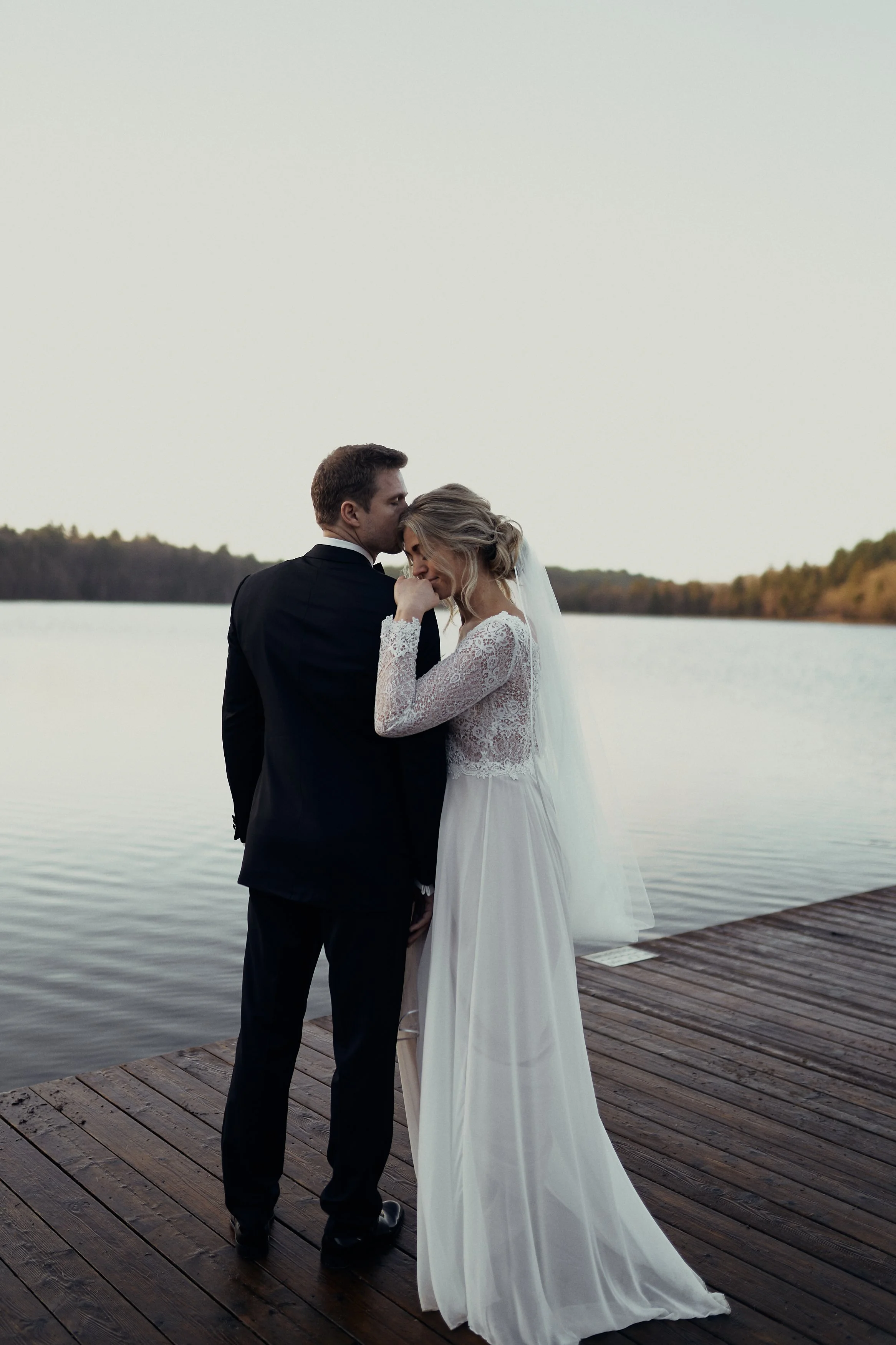 Et nygift står på en træbro ved en Silkeborgsøerne, hvor de kysser. Kvinden har en hvid brudekjole med blondeærmer, og manden har en sort habit. Der er træer i baggrunden. De er blevet gift i 2025.