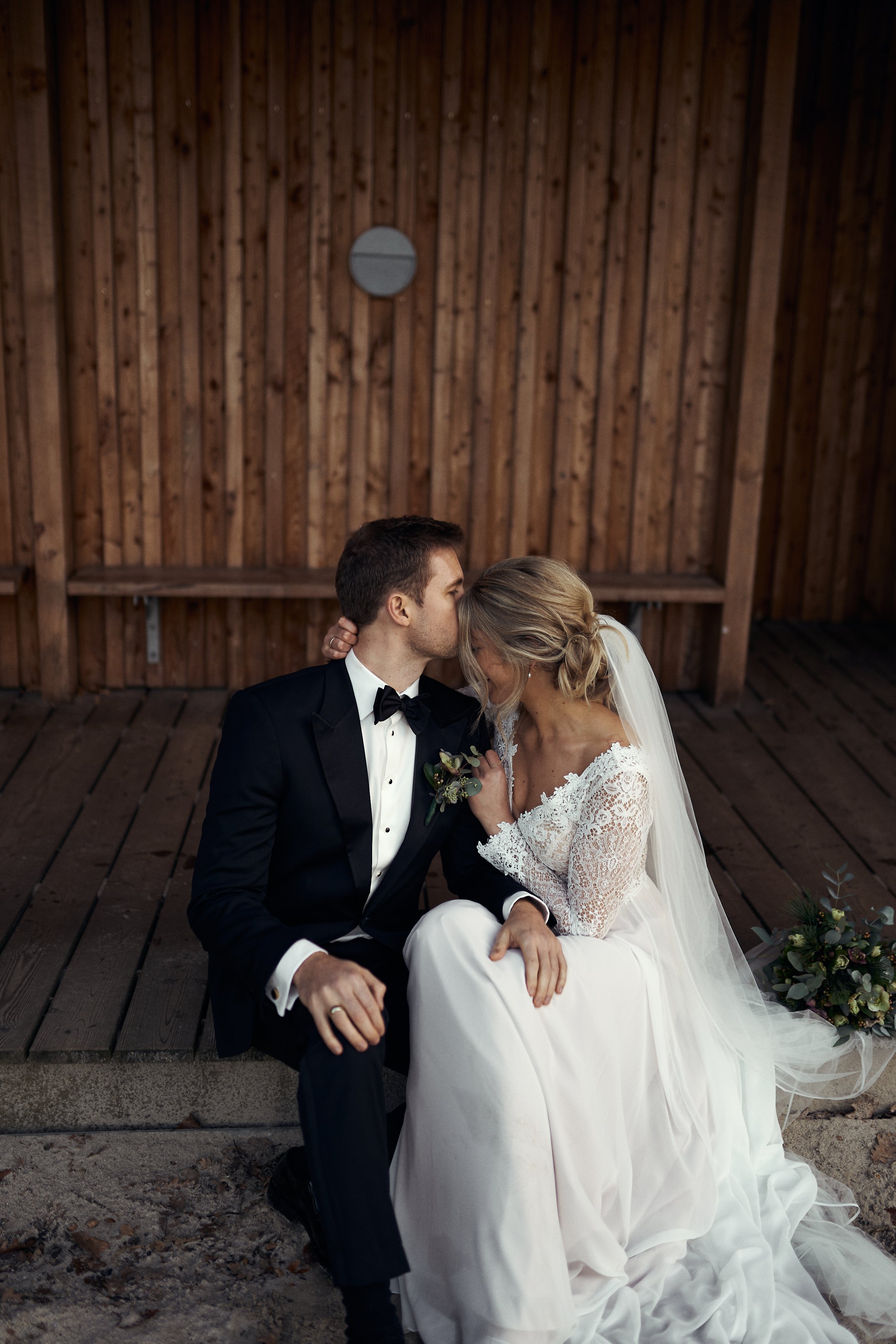 En brud og en brudgom sidder tæt sammen, hvor brudgommen kysser brudens pande på en træbænk foran en trævæg.