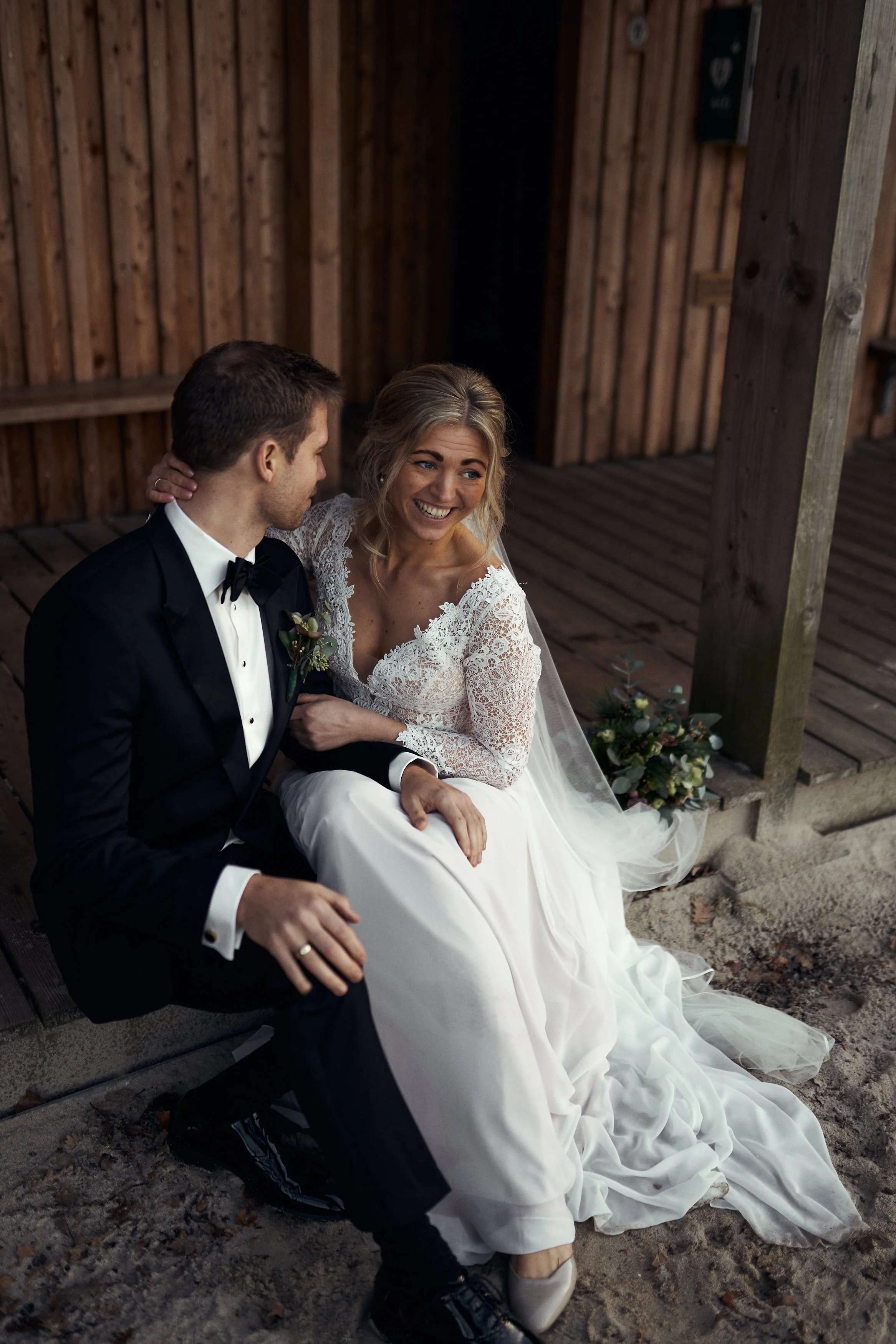 Et brudepar sidder sammen, den mand i sort smoking og den kvinde i hvid brudekjole, smilende, på en træterrasse med trævert væg bagved.