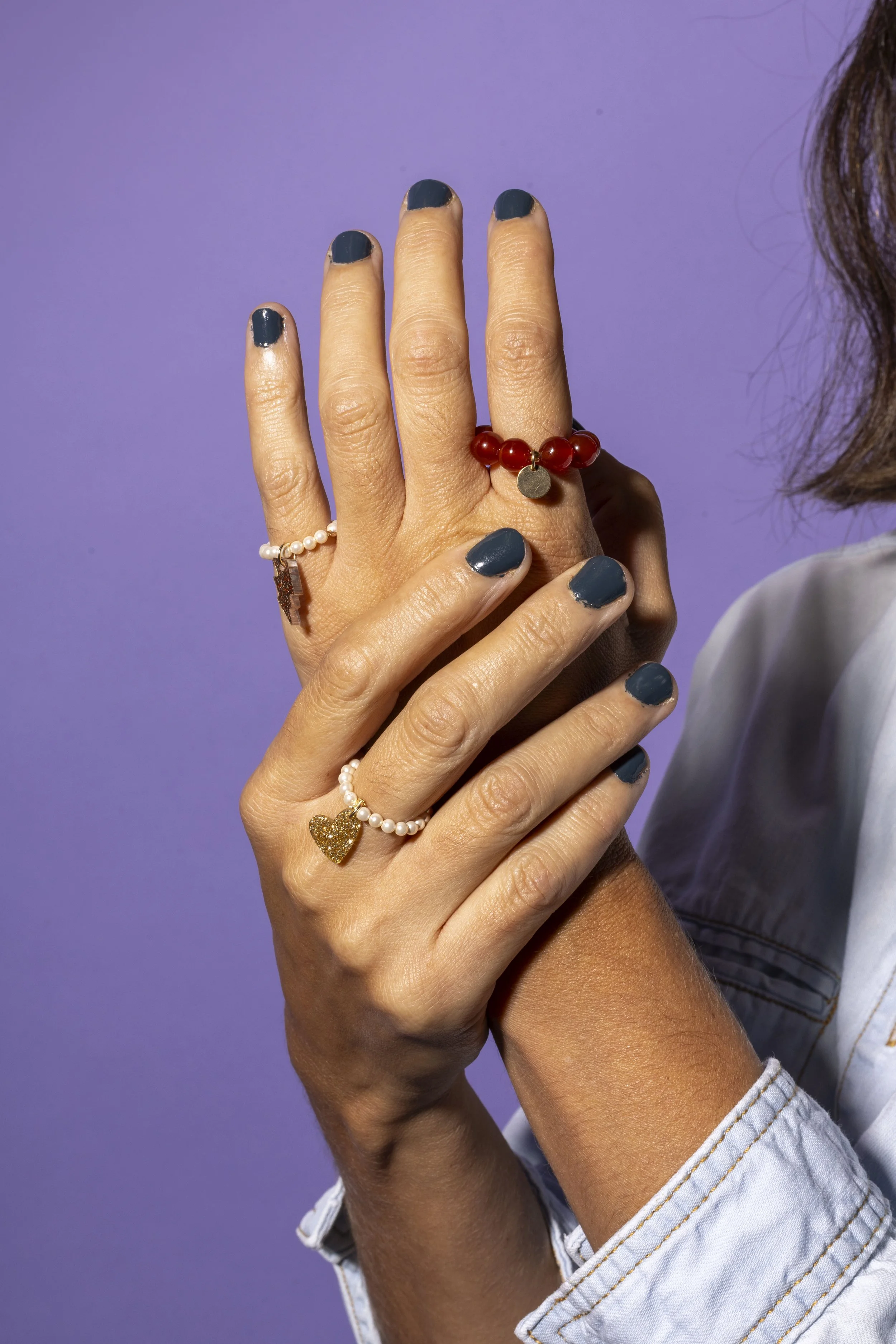 Manos con uñas pintadas de azul, mostrando varios anillos, uno con perlas y corazón dorado, otro con perlas y cruz, y uno con piedras rojas y una pequeña placa, sobre fondo morado.