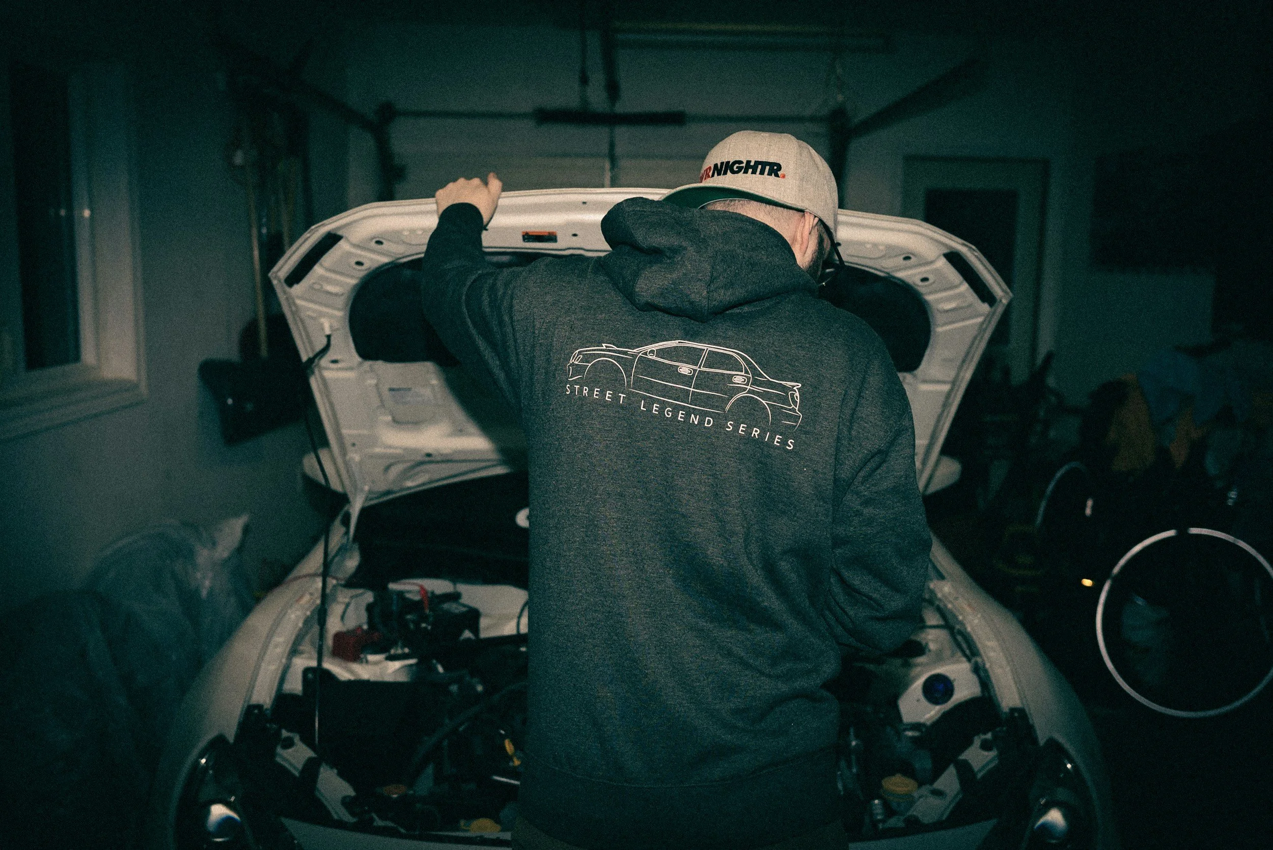 Person working on a car engine in a garage, wearing a dark hoodie with a car outline and 'Street Legend Series' text on the back, and a cap with 'Night' text.