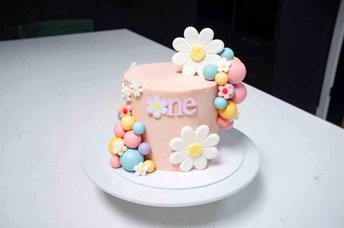 Pink first birthday cake with daisies and rainbow ball decorations