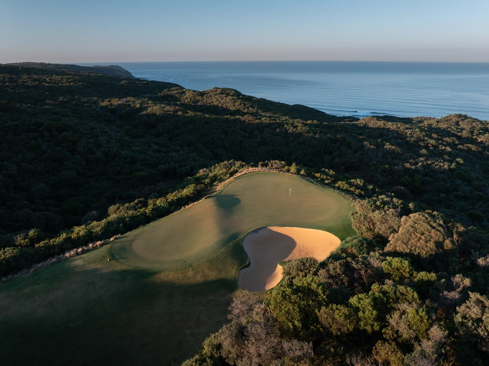 Aerial view of a golf course surrounded by dense trees, with a sandy bunker near the green, overlooking a coastline and ocean in the distance.