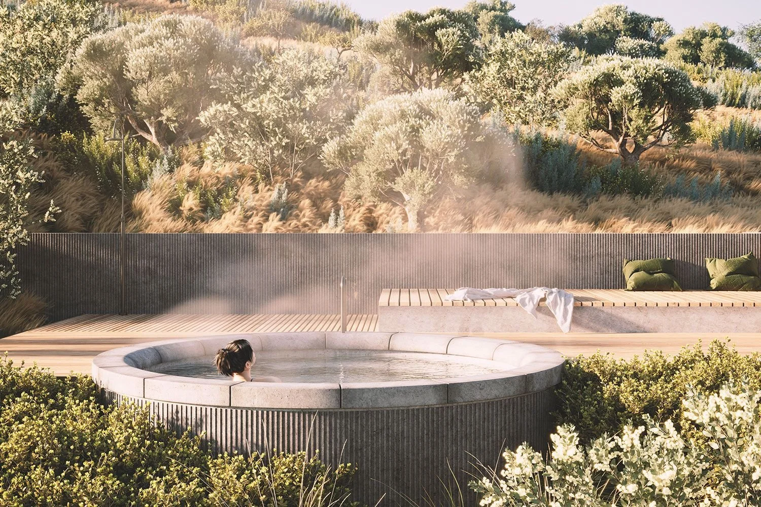 A person sitting in a round hot tub outdoors with steam rising, overlooking a scenic landscape of trees and foliage, with a wooden fence and bench with pillows in the background.