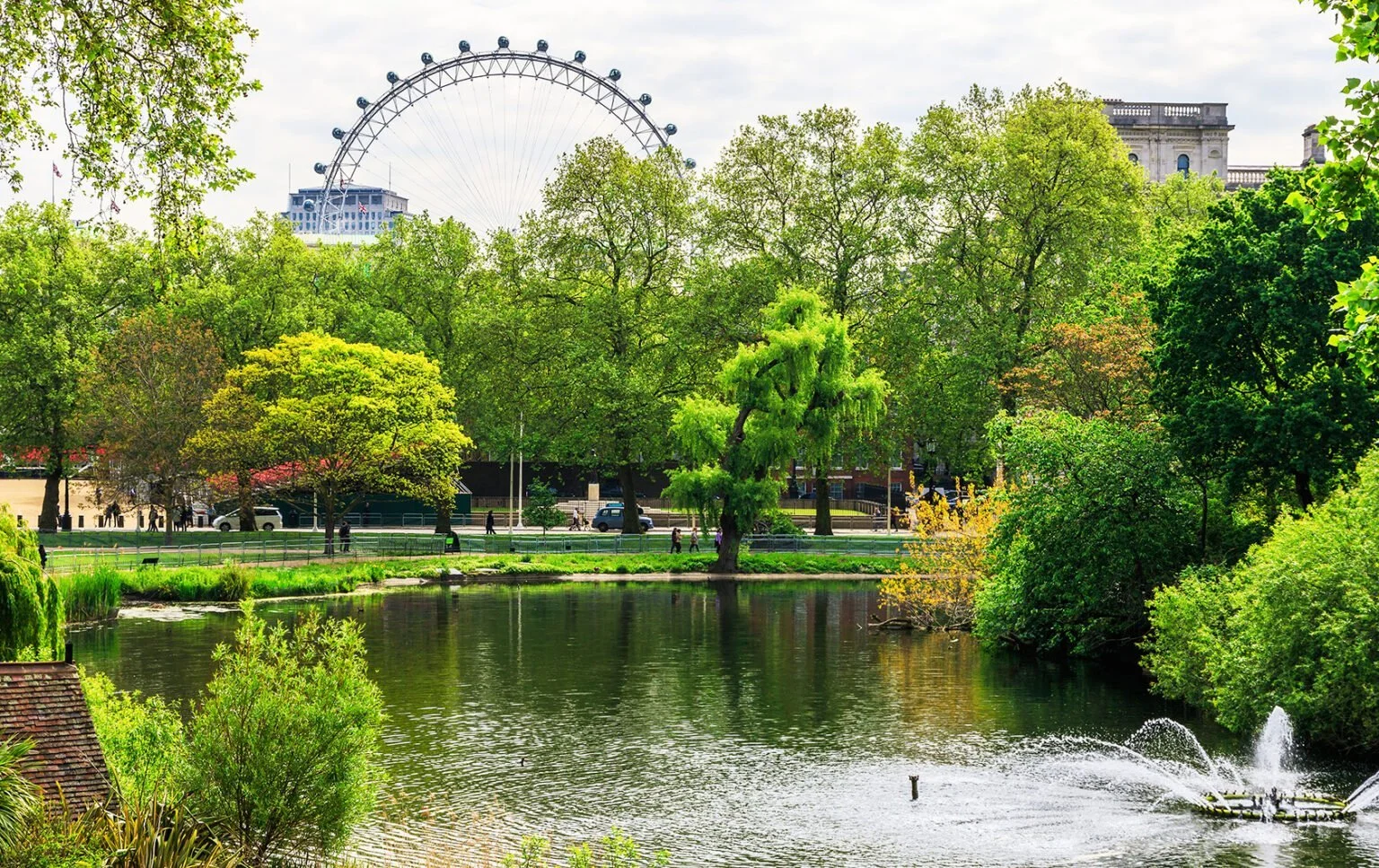 A park with a pond, lush green trees, and a large Ferris wheel in the background.