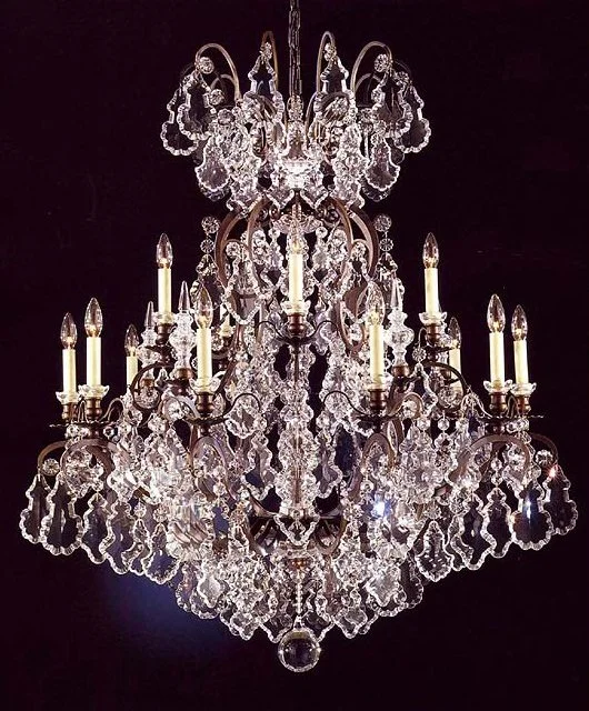 A large, ornate crystal chandelier with multiple lit candles, intricate hanging crystals, and a dark background.
