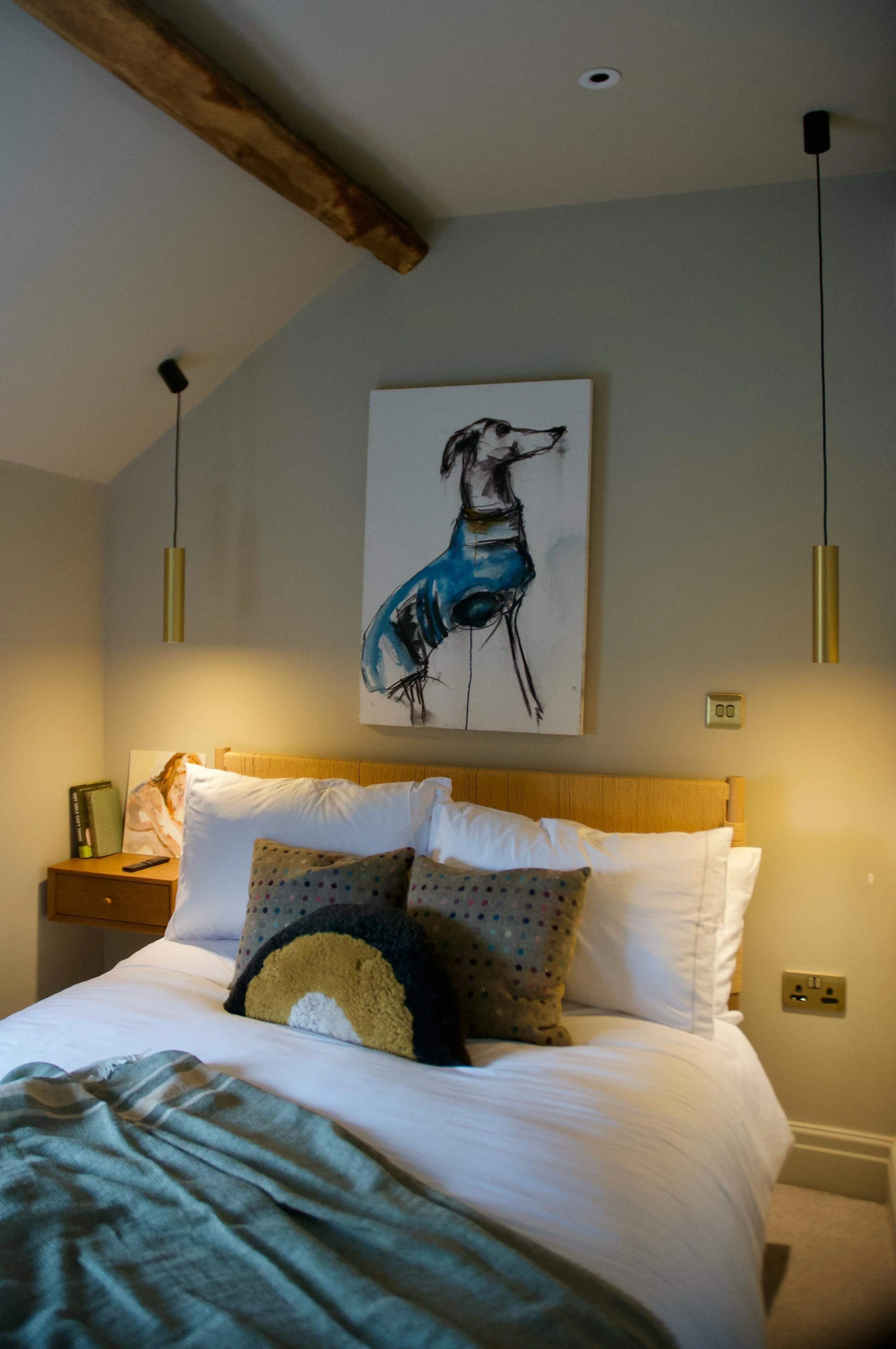 A cozy bedroom with a bed featuring white sheets and pillows, a wooden headboard, and decorative pillows. A bedside table holds books and a framed photo. Above the bed is an abstract painting of a dog in black, blue, and yellow. Two hanging pendant lights are on either side of the painting. The walls are painted in neutral colors, and there is a wooden ceiling beam.