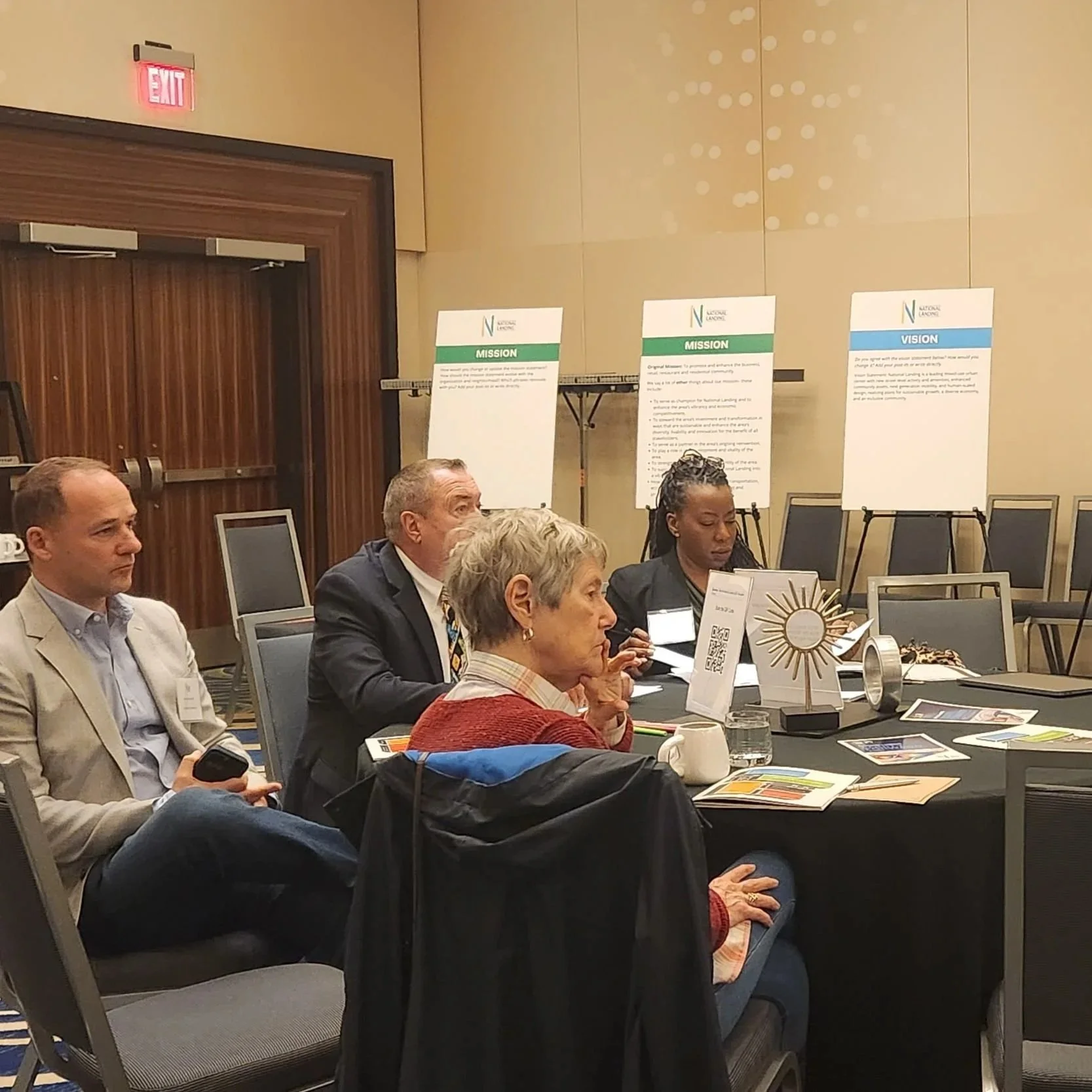 People seated at round tables during a workshop, with mission, vision, and values posters displayed on easels in the background.