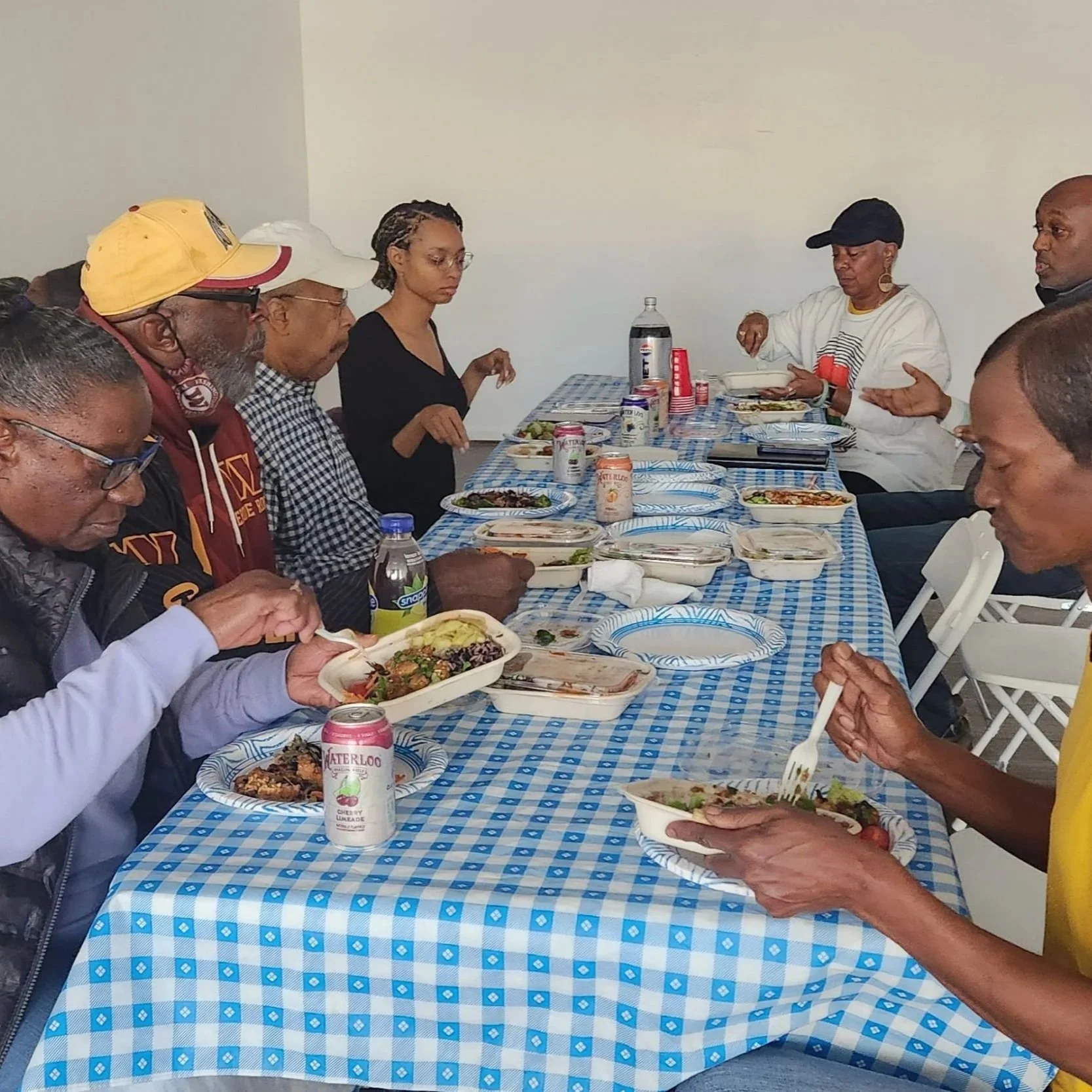 A group of Skyland Terrace residents sit around a long table with a blue and white checkered tablecloth, sharing a meal served in individual paper containers.