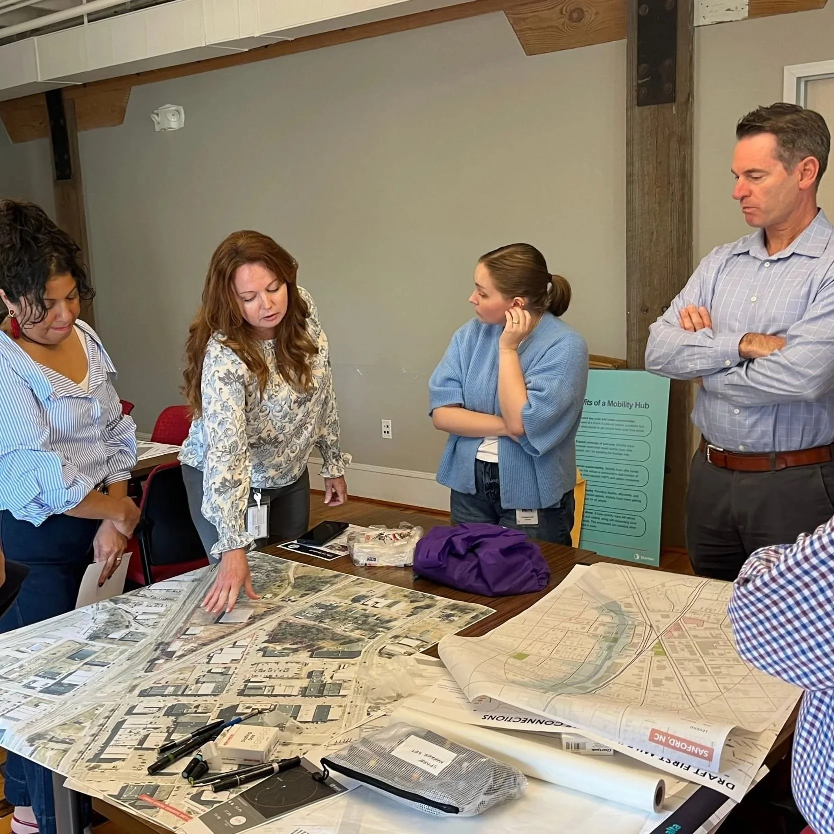 A small group of five people engaged in a focused discussion, leaning over a table to review and examine several large maps or planning documents.