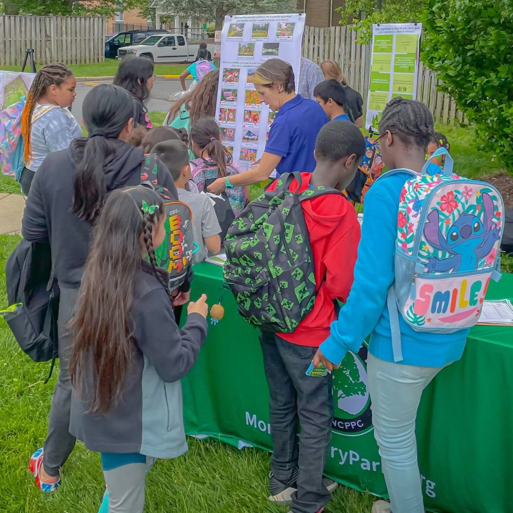 Community members and children gather outdoors around informational tables and display boards during a local engagement event.