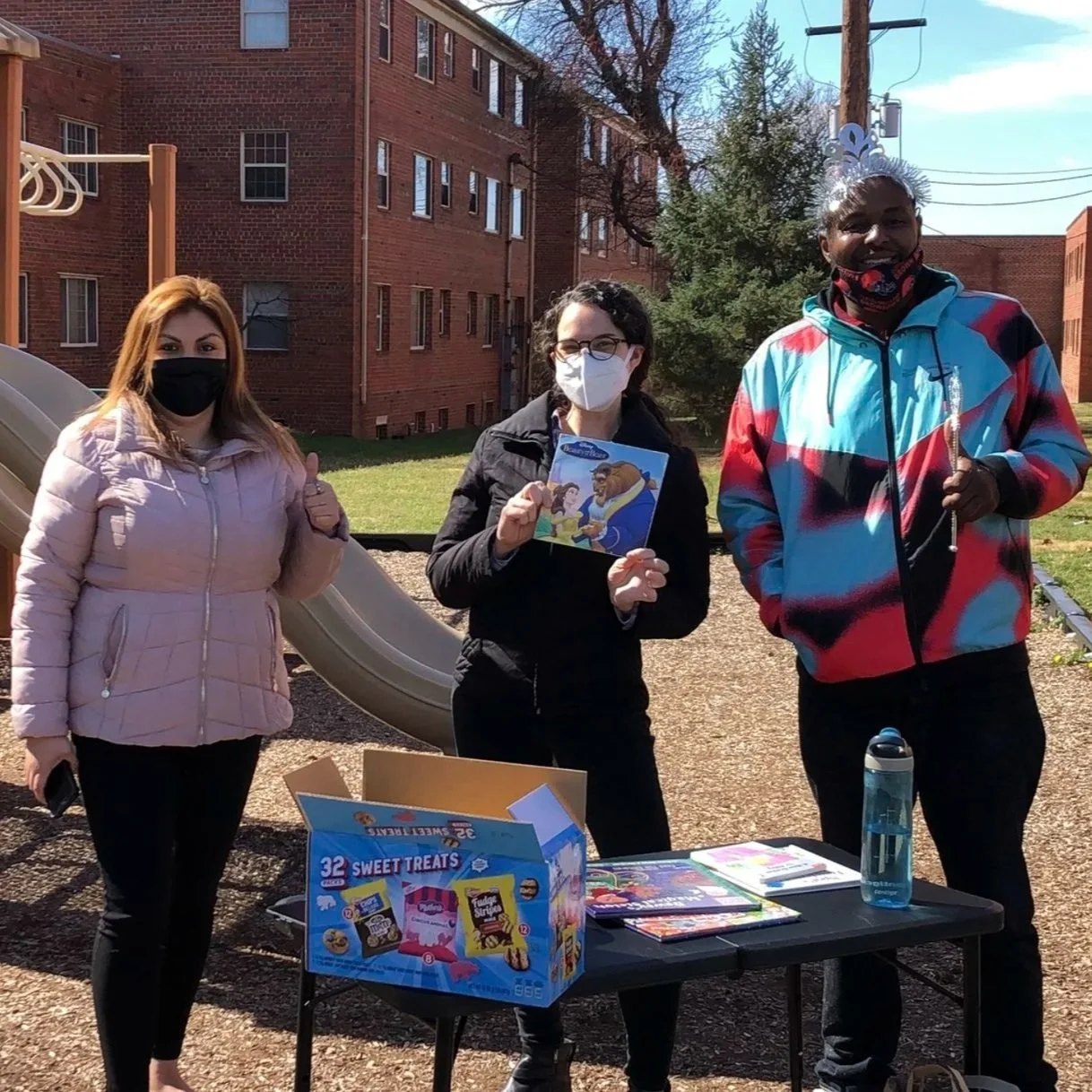 Small outdoor information table set up near a playground and brick apartment buildings, with three women standing behind the table interacting with a passerby.