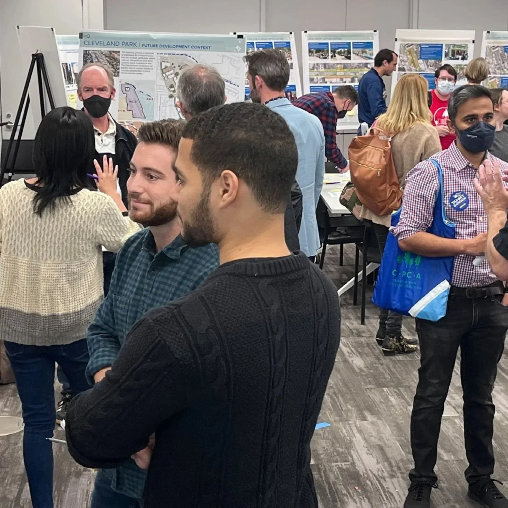Crowded community engagement event where people stand in small groups to read and discuss informational posters on easels.