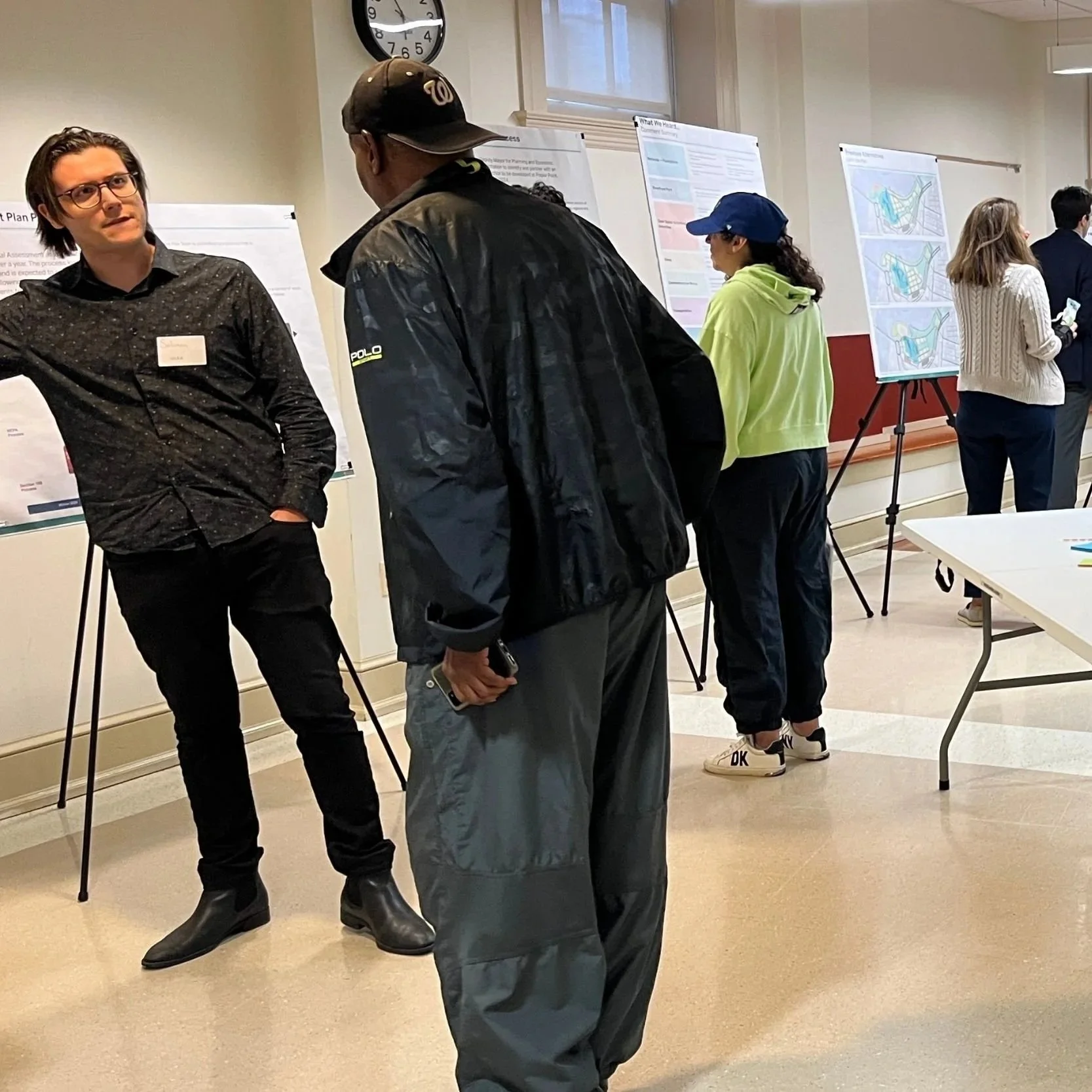 Indoor scene of a gathering with several people standing and looking at white posters mounted on easels; a man in a black shirt speaks to another attendee near a central white table.