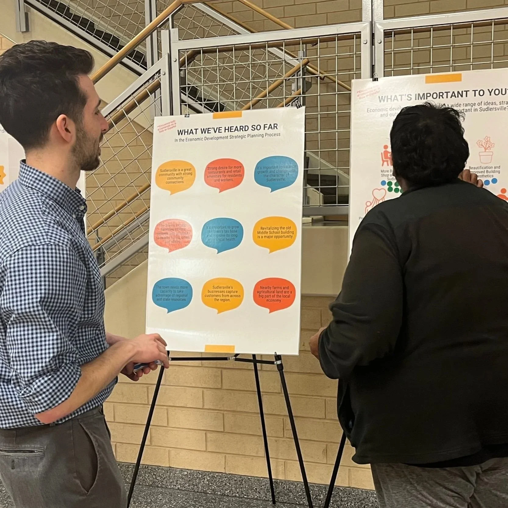 Two people stand indoors reviewing community feedback posters on easels, including boards titled “What We’ve Heard So Far” and “What’s Important to You?”, with sticky notes and other displays visible at a public engagement event.