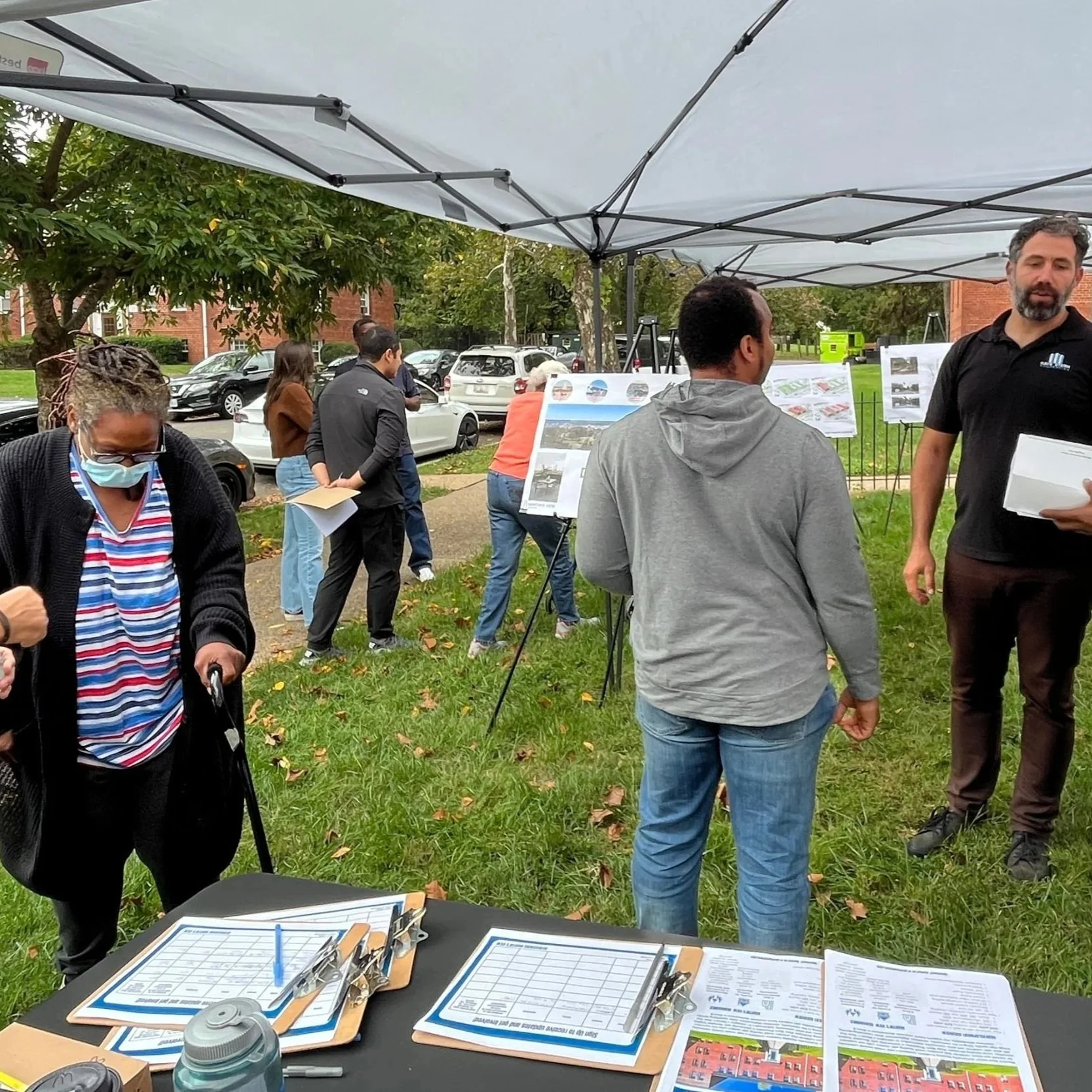 Community members and staff gather under outdoor canopy tents at a neighborhood engagement event, viewing display boards, talking with facilitators, and signing in at a table with clipboards.