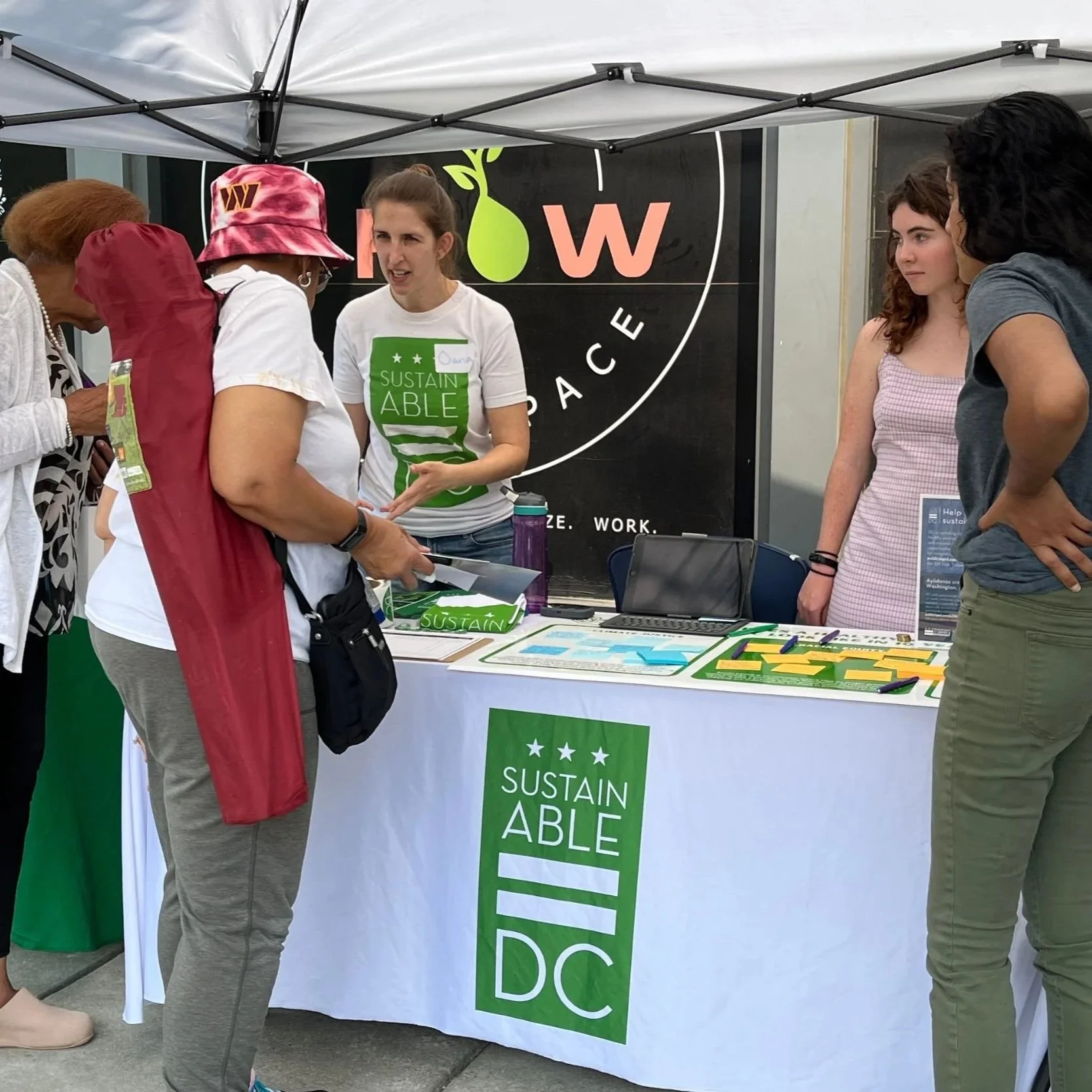 Two staff members at the "Sustainable DC" booth under a white tent engaging with two women attendees at an outdoor fair.