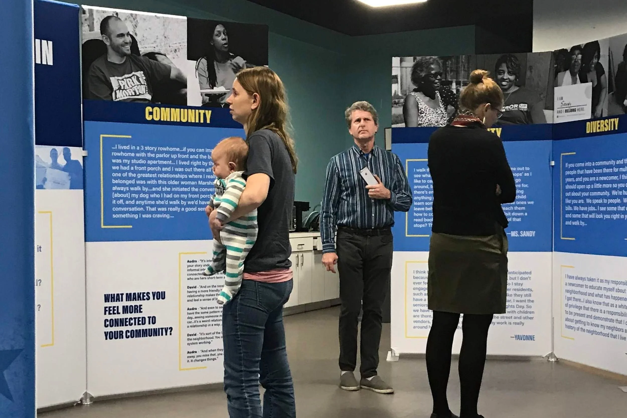 Visitors stand inside a community exhibit, reading blue-and-white panels about community and diversity; one person holds a baby while others view the displays.