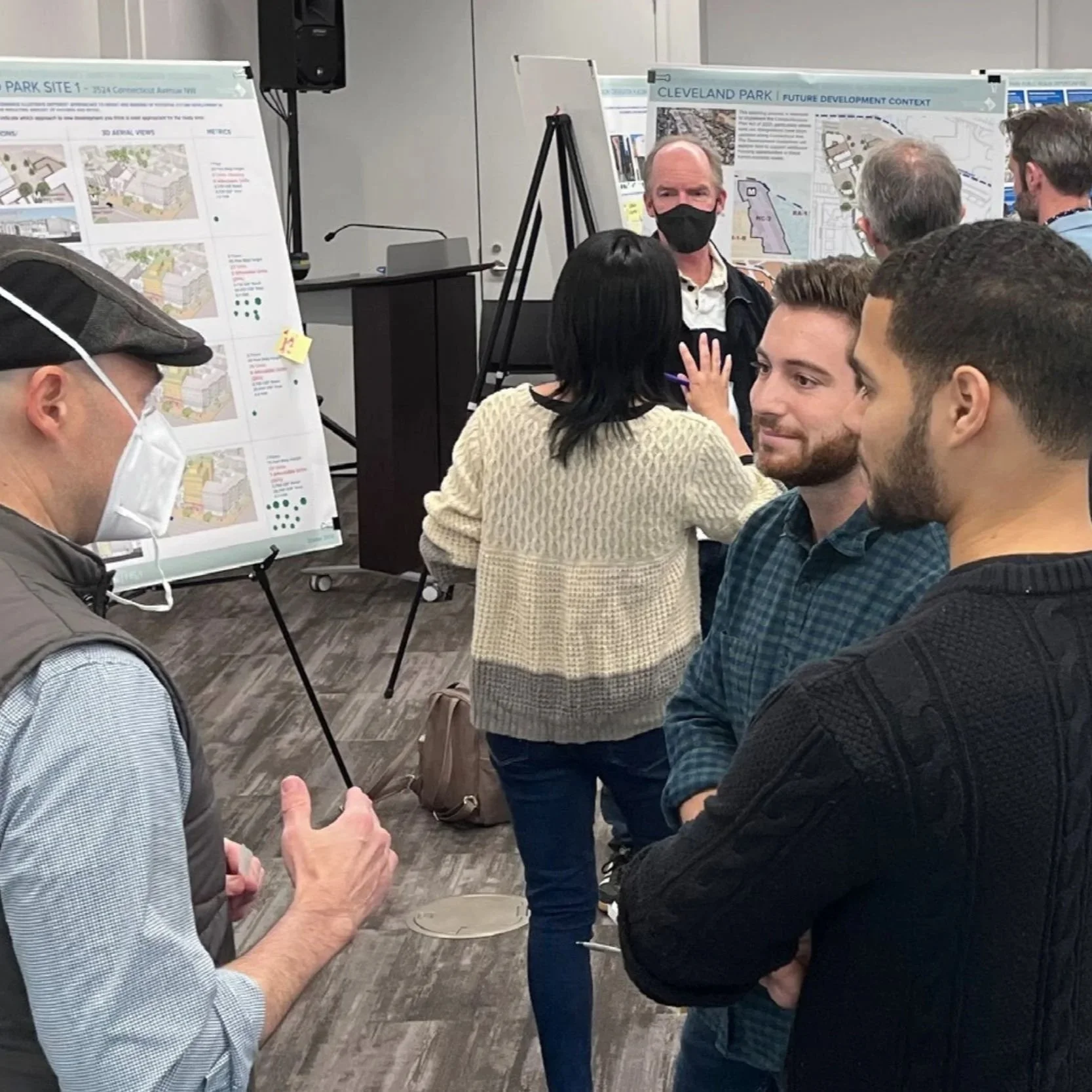 Crowded community engagement event where people stand in small groups to read and discuss informational posters on easels.