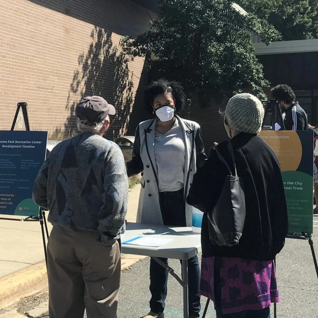 Attendees and a staff member gathered outdoors at an informational table near a building entrance, engaging in discussion.