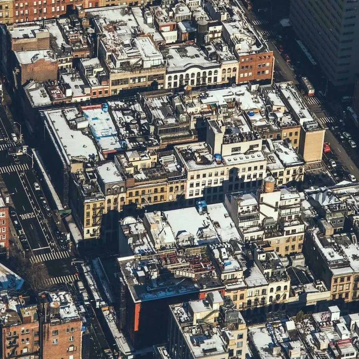 Aerial view of dense urban neighborhood rooftops in Harlem, featuring mixed architecture, snow-dusted roofs, water towers, and busy streets below, with clear blue sky above.