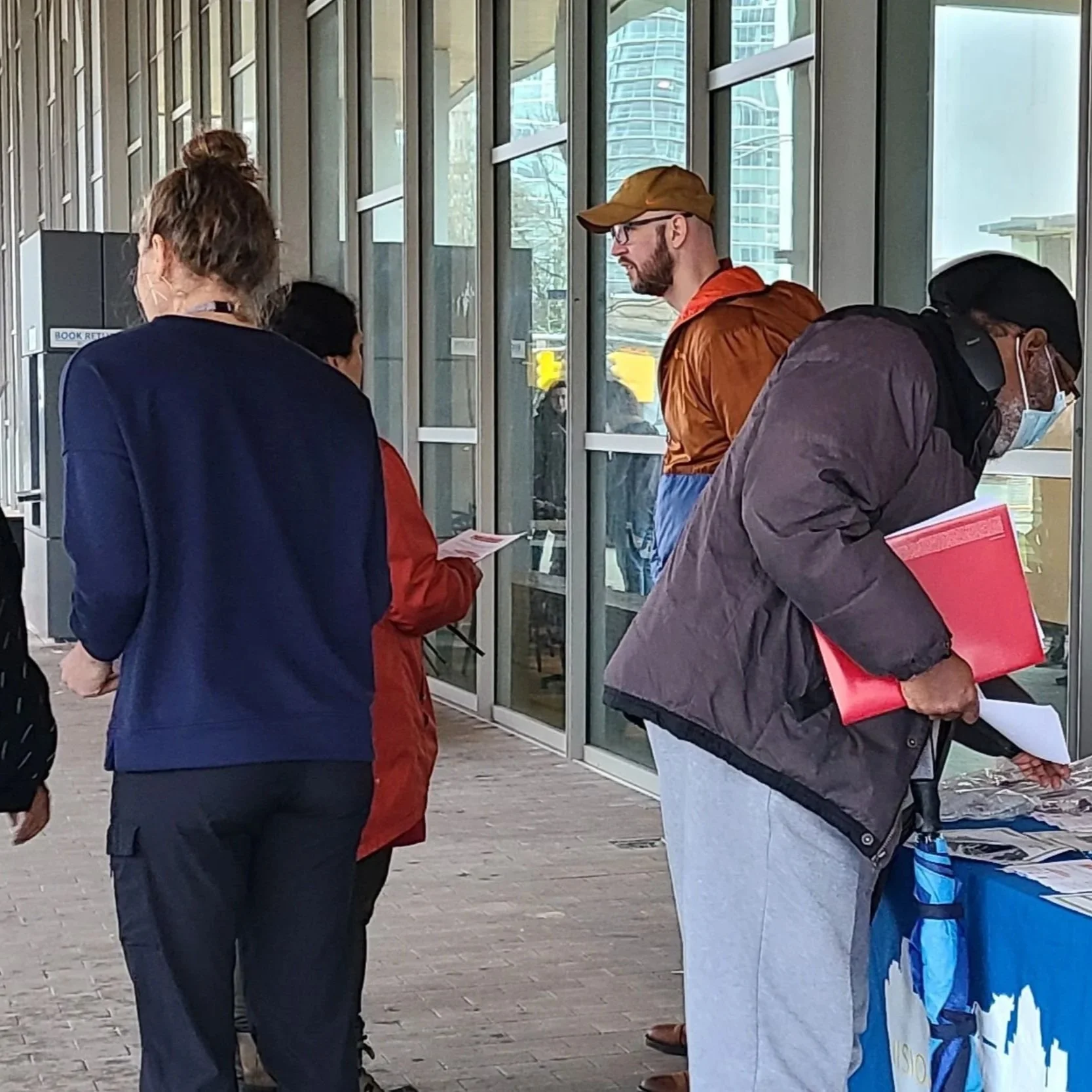 People gathered outside a building at an informational table covered with pamphlets and materials, with people engaging in conversation on a cool day.