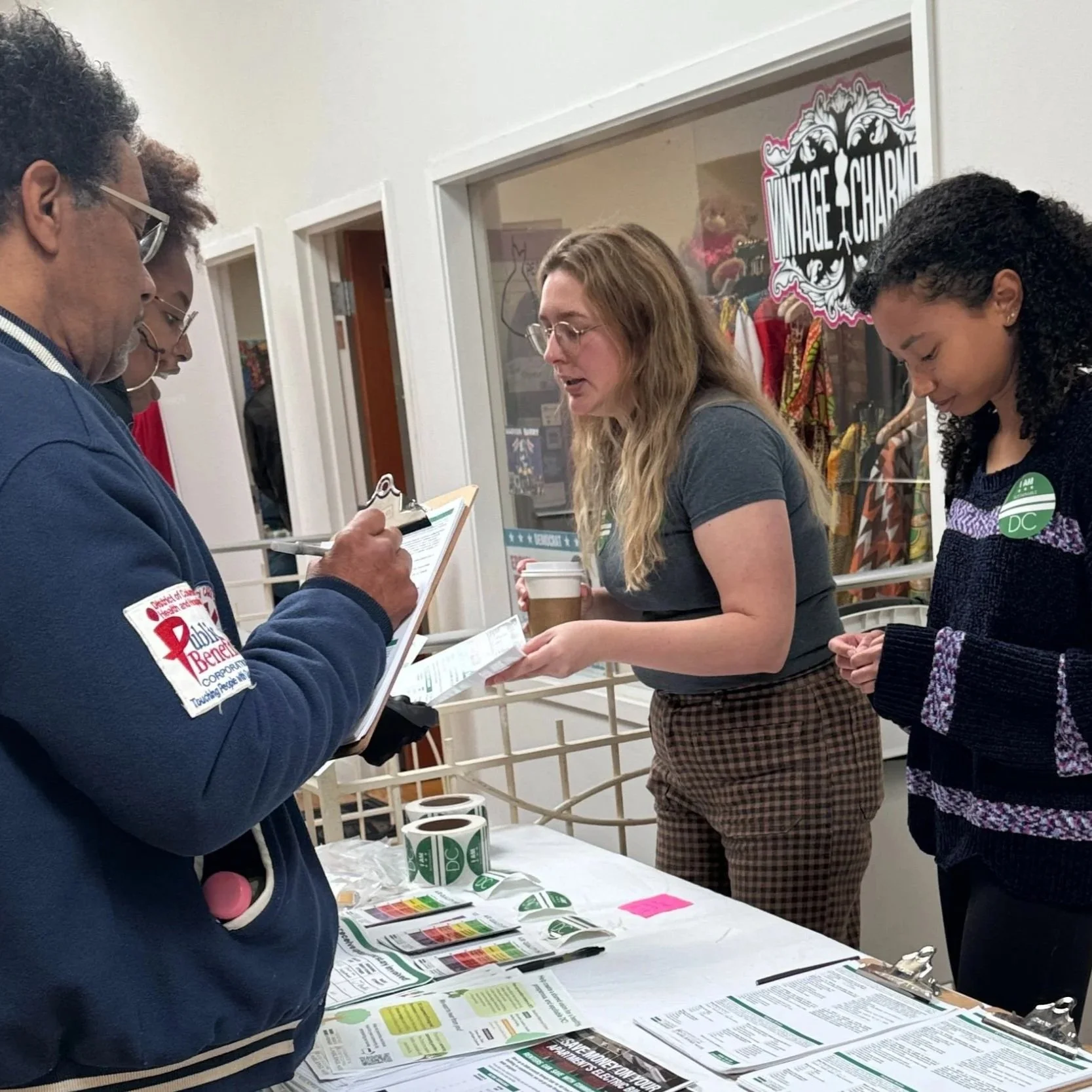 Two people standing indoors at a table with a display board titled 'Share Your Ideas on Climate Change in Your Community,' one writing on a clipboard and the other looking at materials on the table.