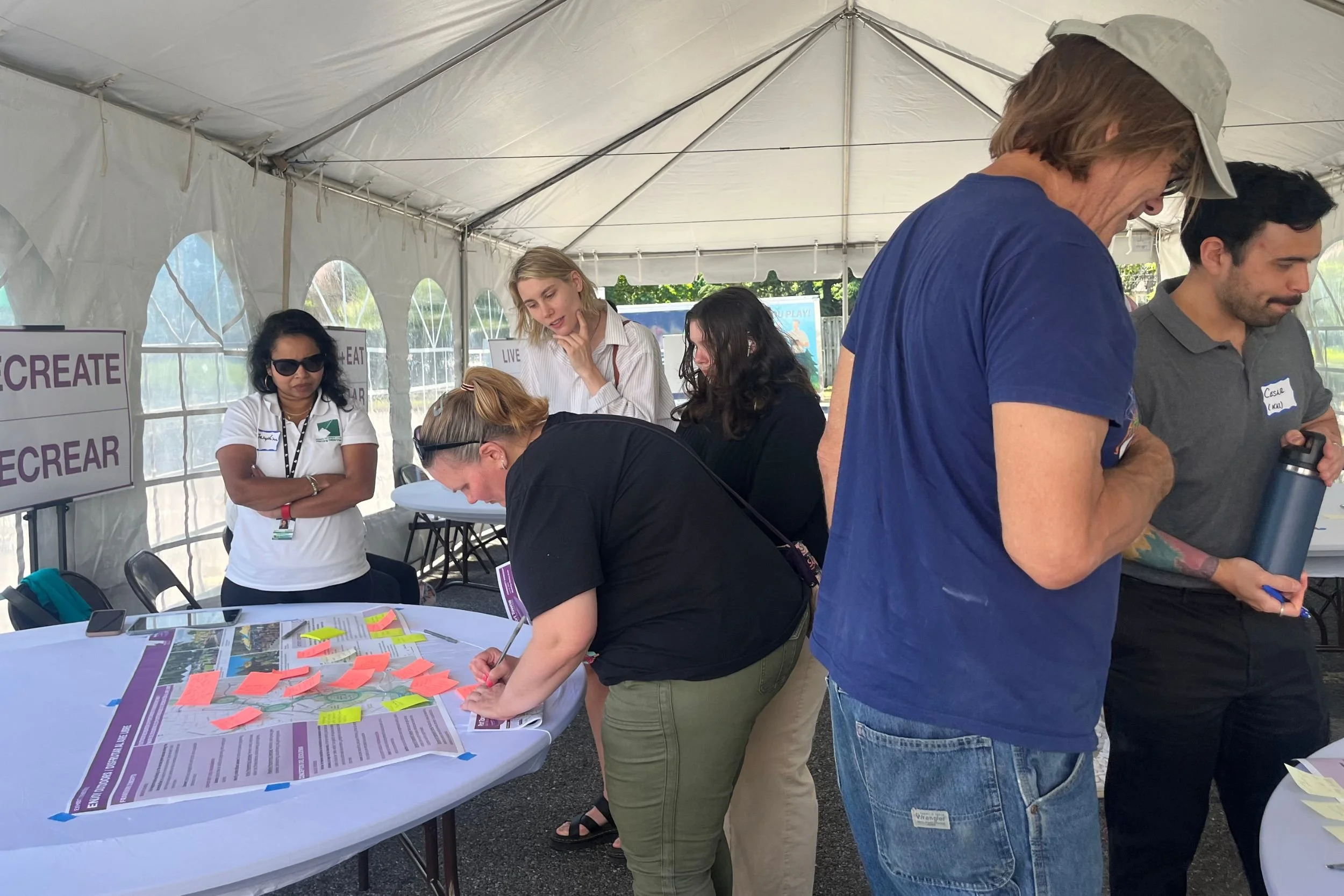 Workshop group standing around a table covered in sticky notes and maps, collaborating on a project under a white event tent.