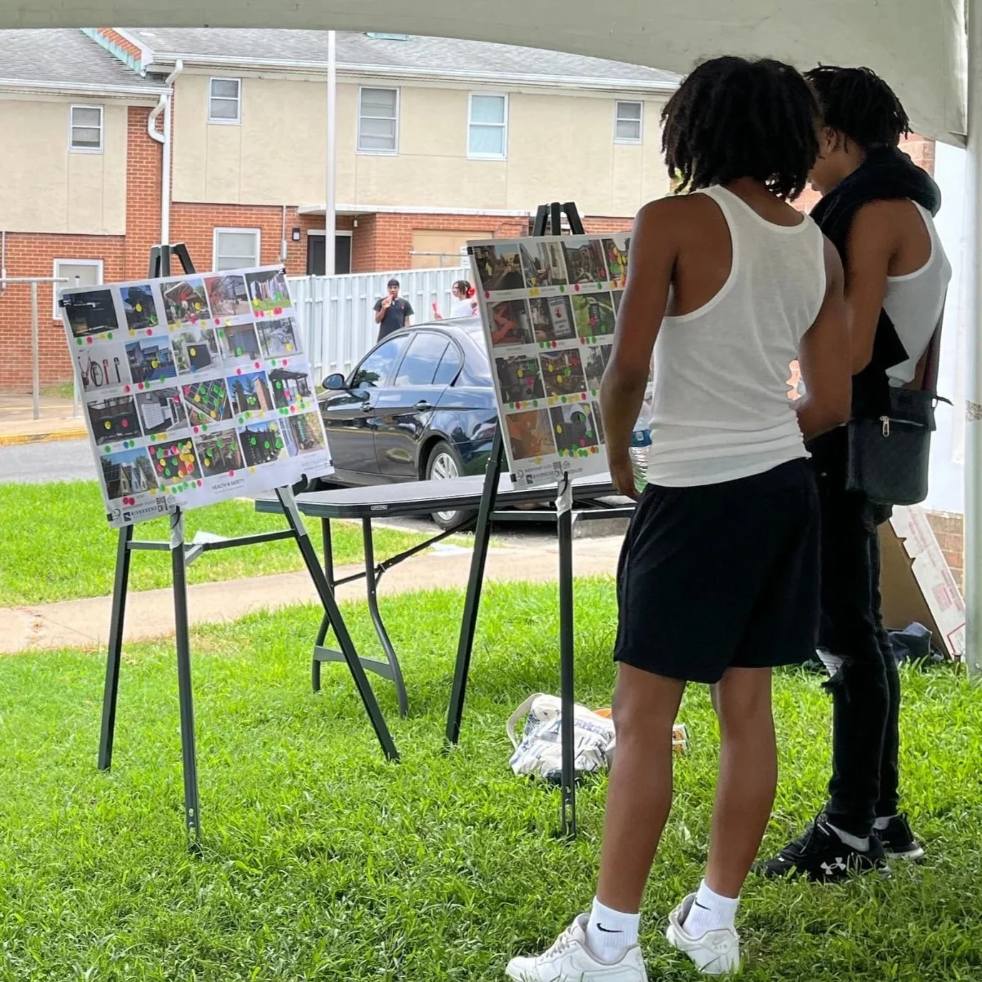 Attendees at an outdoor community engagement session gathered under a tent on the grass, reviewing informational posters on easels near residential buildings.