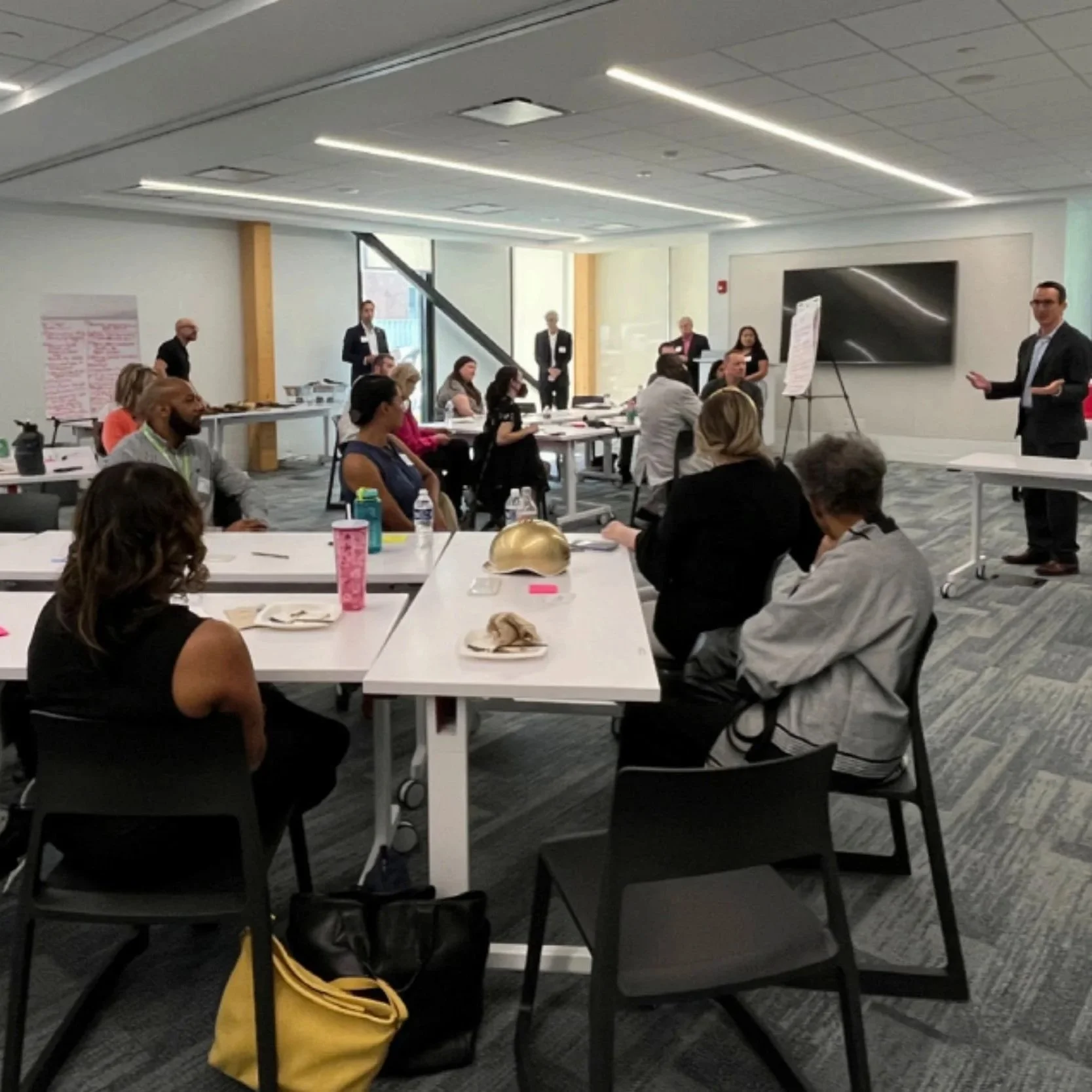 Large group attending a meeting in a contemporary, brightly lit room with a speaker standing at the front and attendees seated in rows.