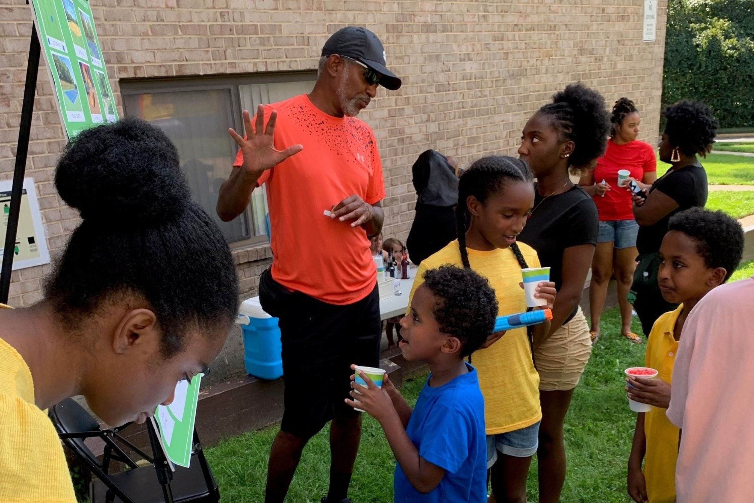 Outdoor community event where adults and children are talking and viewing information presented on a display board near a brick building.