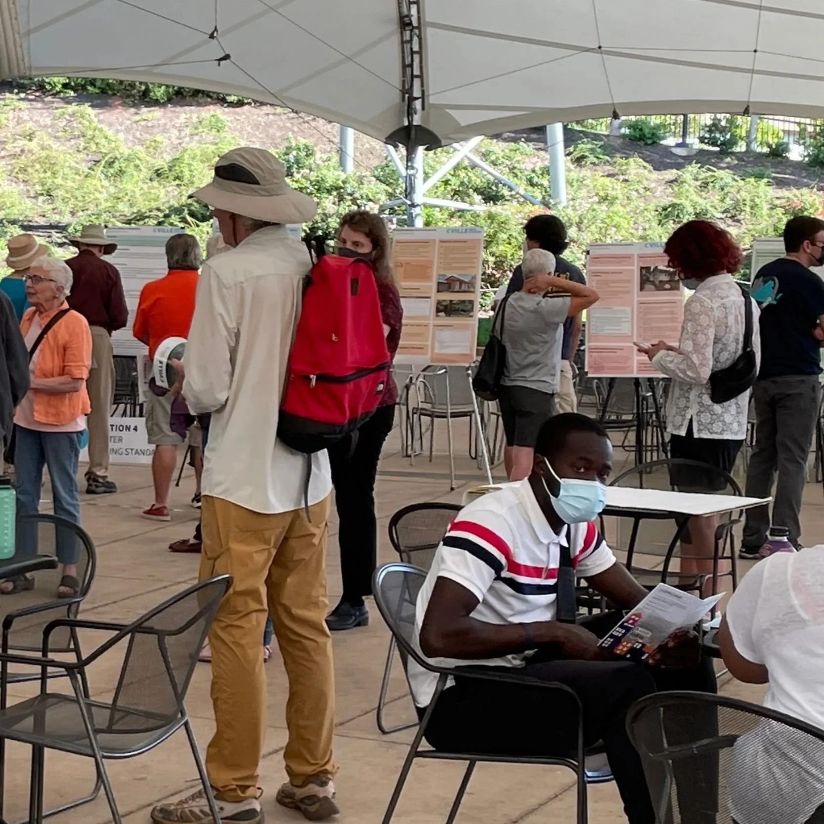 Group of people gathered under a large outdoor canopy, viewing informational posters and engaging in discussions at a community event