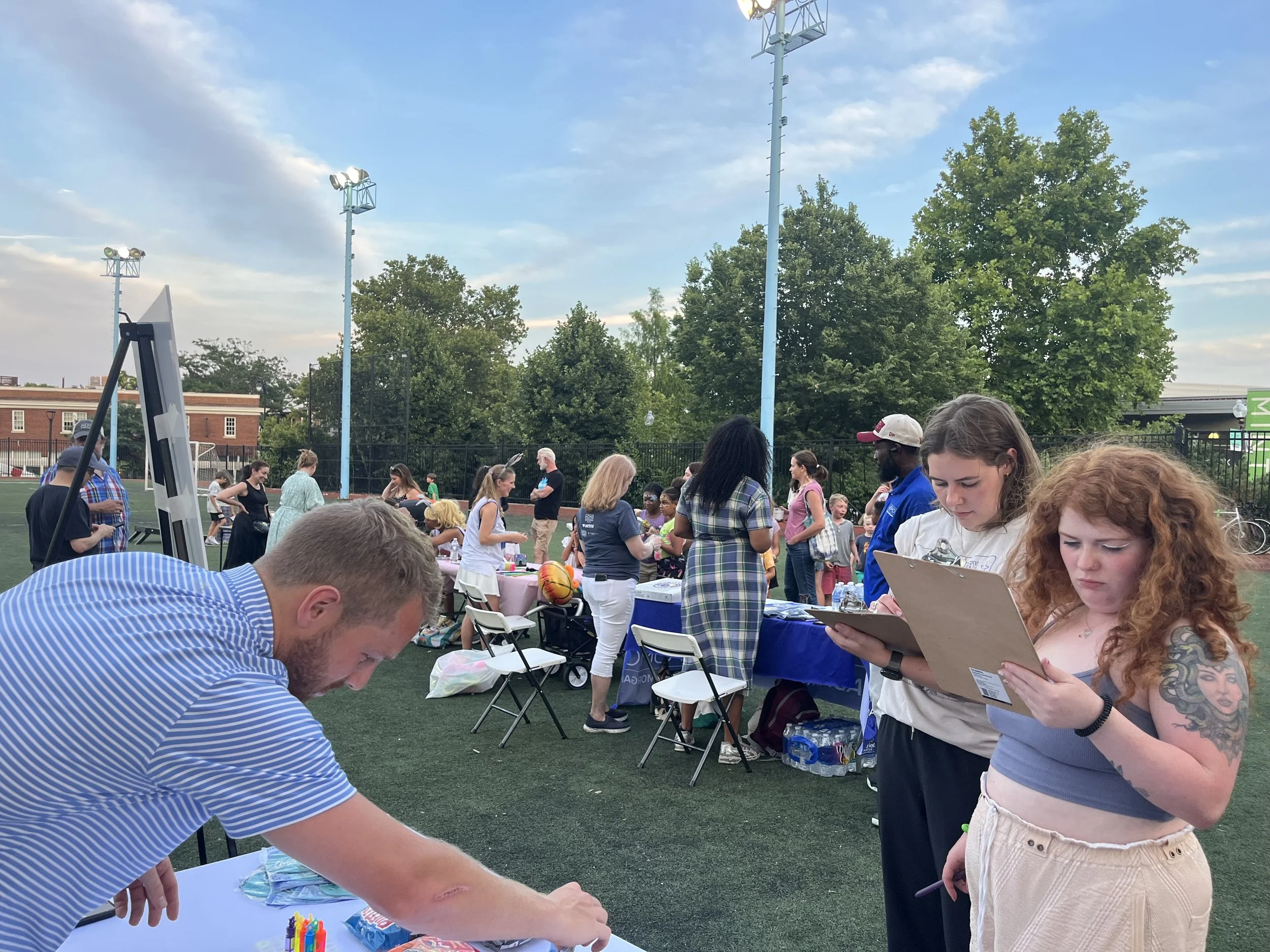 People gather at an outdoor event with tables set up on a field. Some community members fill out forms while others talk and interact near informational exhibits.