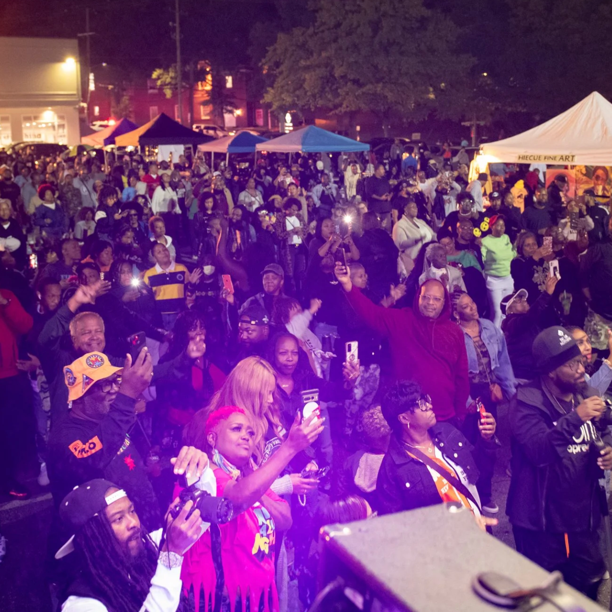 A large, diverse crowd gathers at night for an outdoor community festival or block party, featuring vendor tents, stage lighting, and many attendees holding up phones to capture the event.
