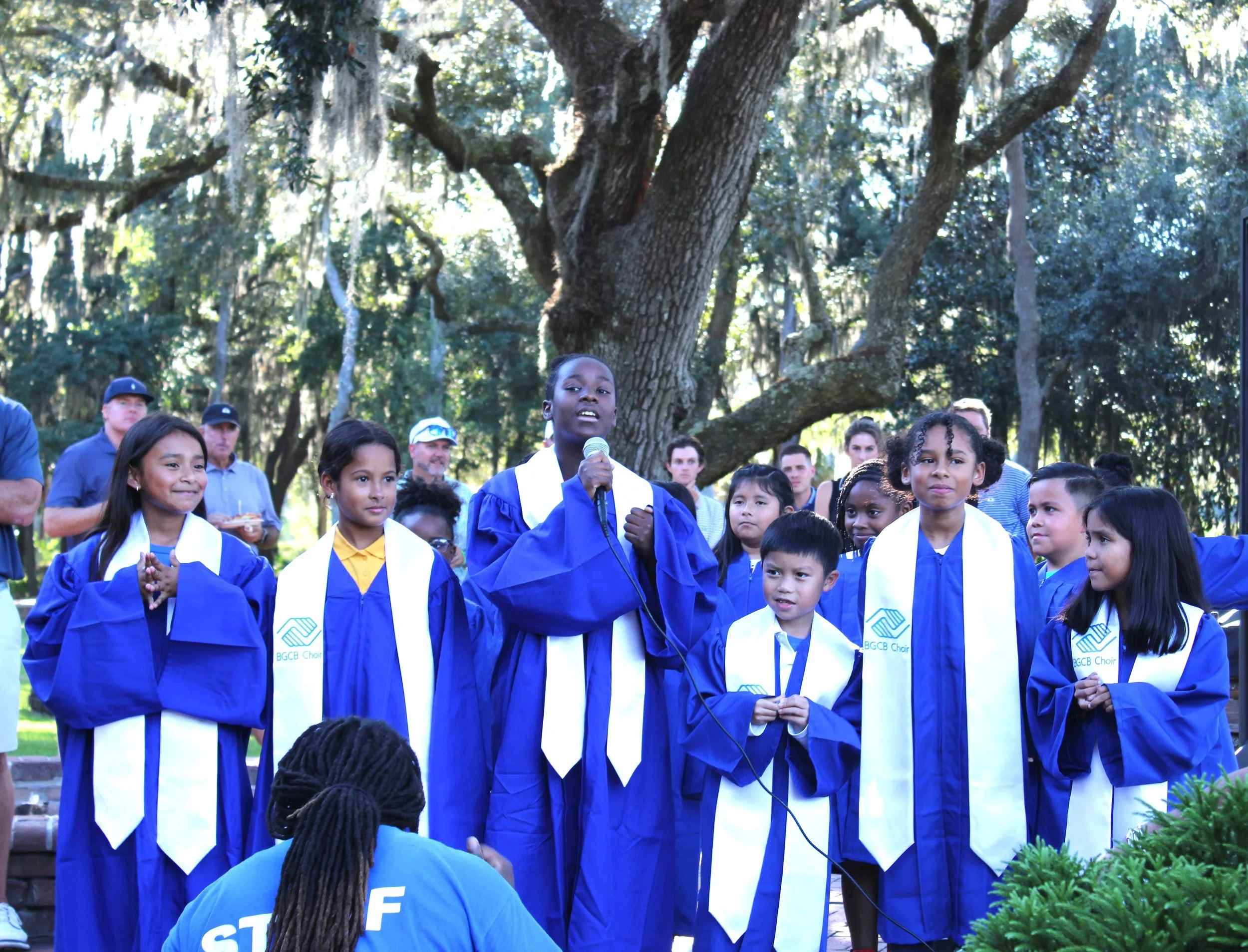 A group of children in blue and white graduation gowns standing outdoors under large trees, with some adults in the background, during a graduation ceremony. One girl is speaking into a microphone.