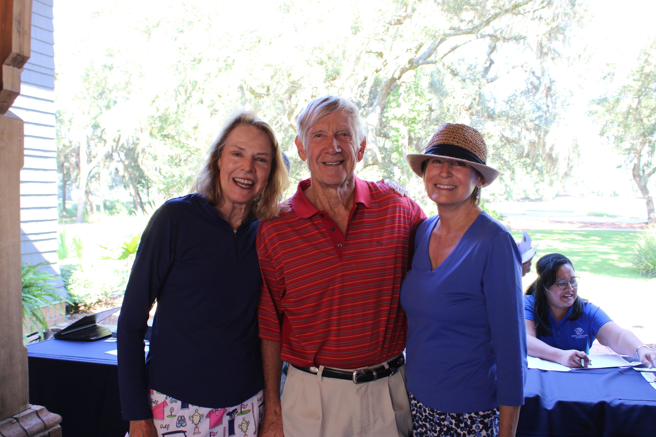 Three elderly adults smiling and posing outdoors, with a background of trees and a partially visible woman sitting at a table, wearing a blue shirt and glasses.