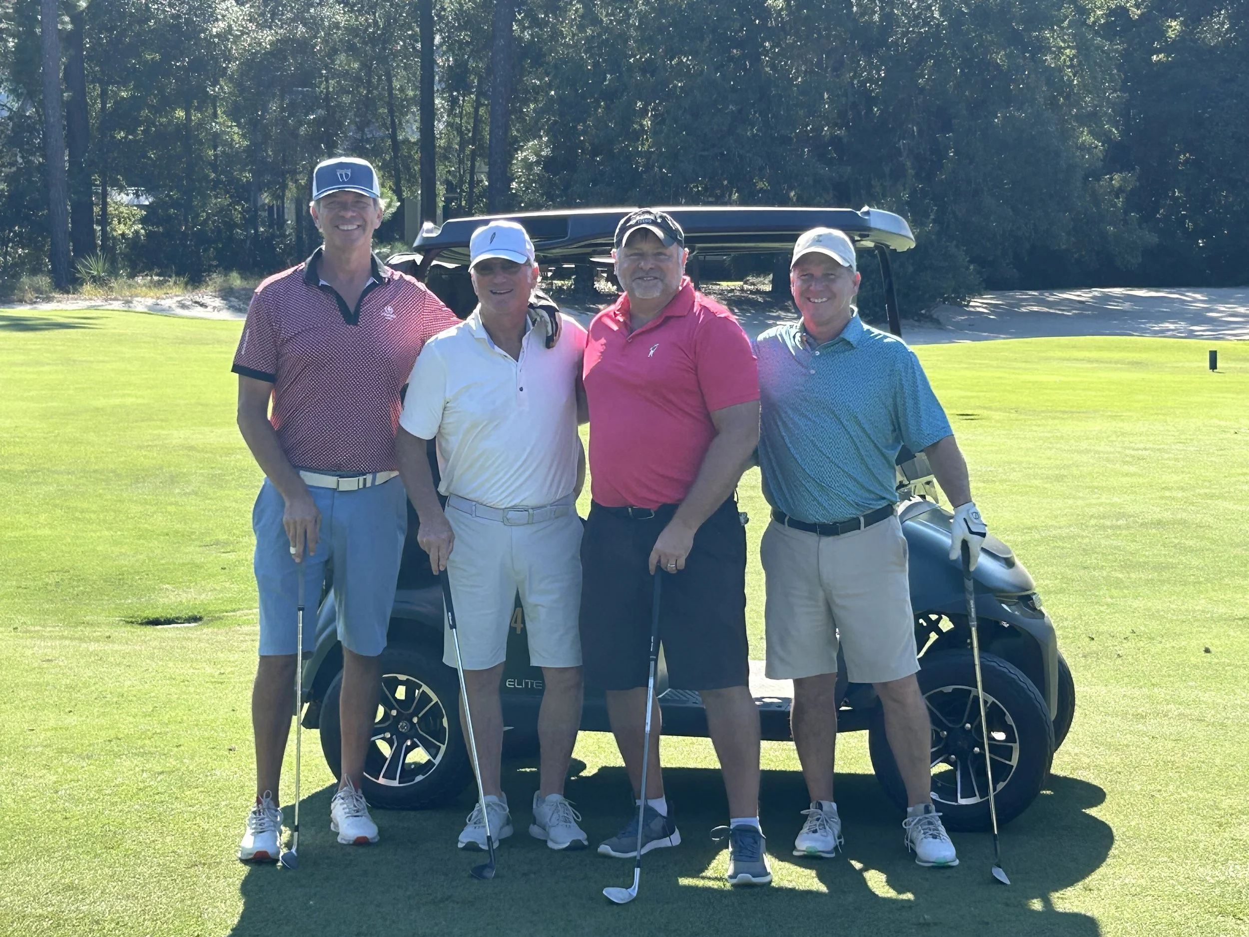 A group of four men standing on a golf course, smiling, with golf clubs in hand and a golf cart behind them.