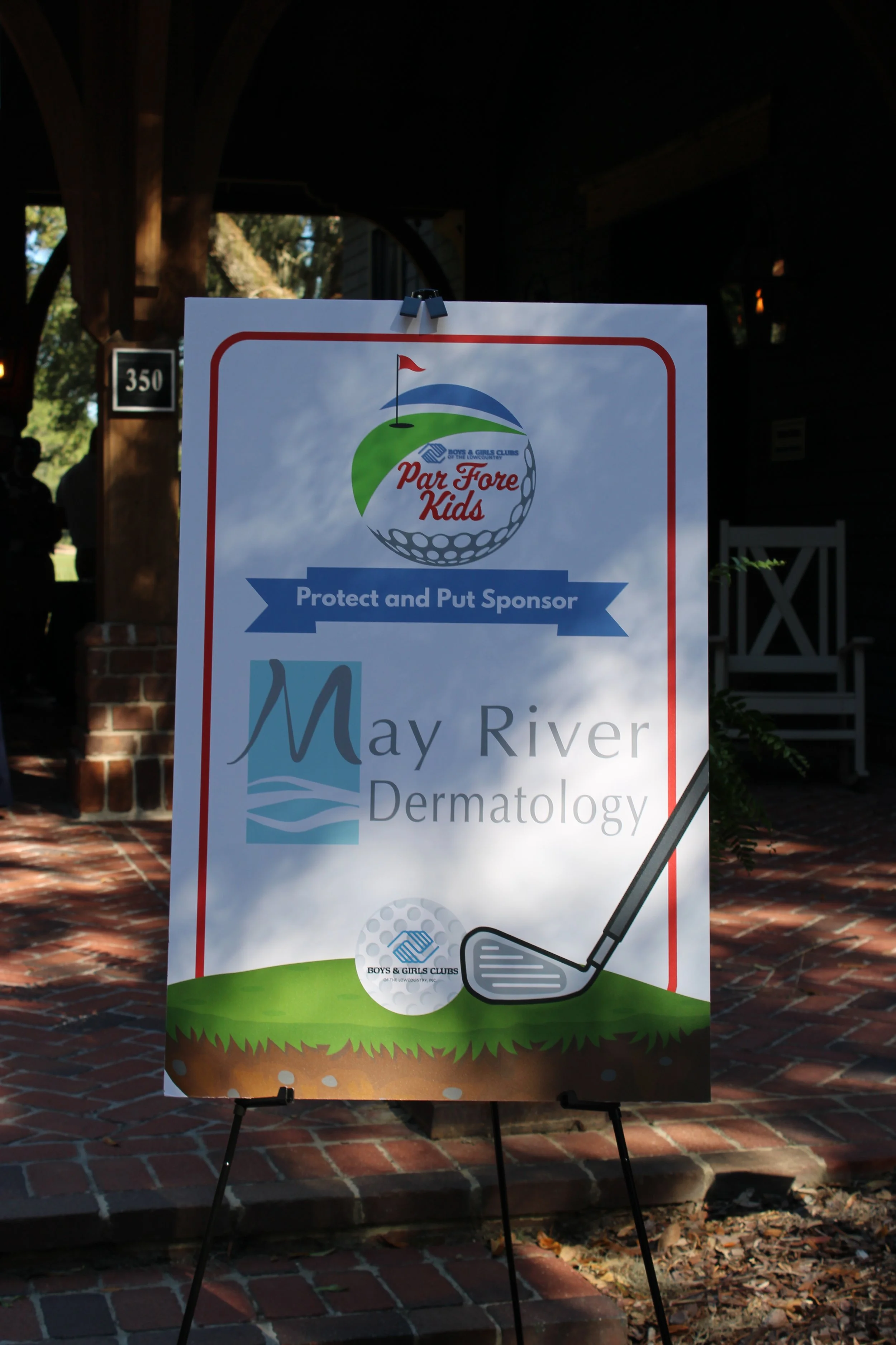 A sign promoting a golf event called 'Par Fore Kids' by the Boys & Girls Clubs of the Lower Country, featuring a golf ball with a golf club, and a background with trees and a building.