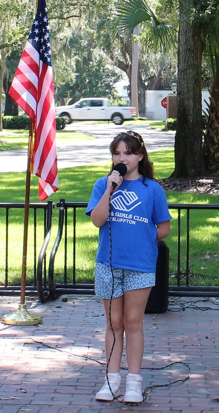 A young girl in a blue T-shirt and shorts speaking into a microphone outdoors during an event, with an American flag and trees in the background.