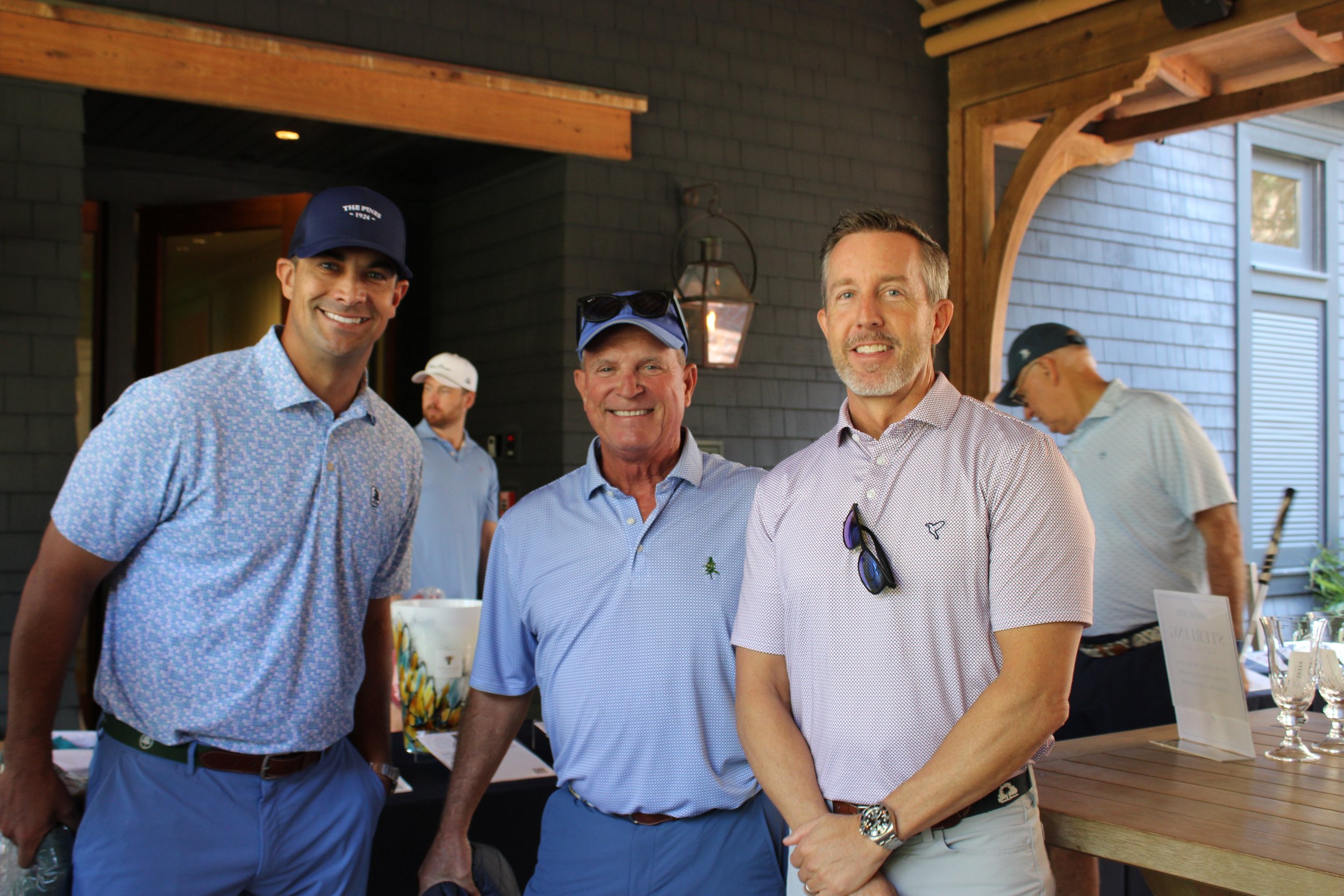 Three men smiling and standing close together at a social gathering or outdoor event. One is wearing a light blue patterned shirt and a navy cap, another in a blue shirt with sunglasses hanging from his collar, and the third in a light patterned shir